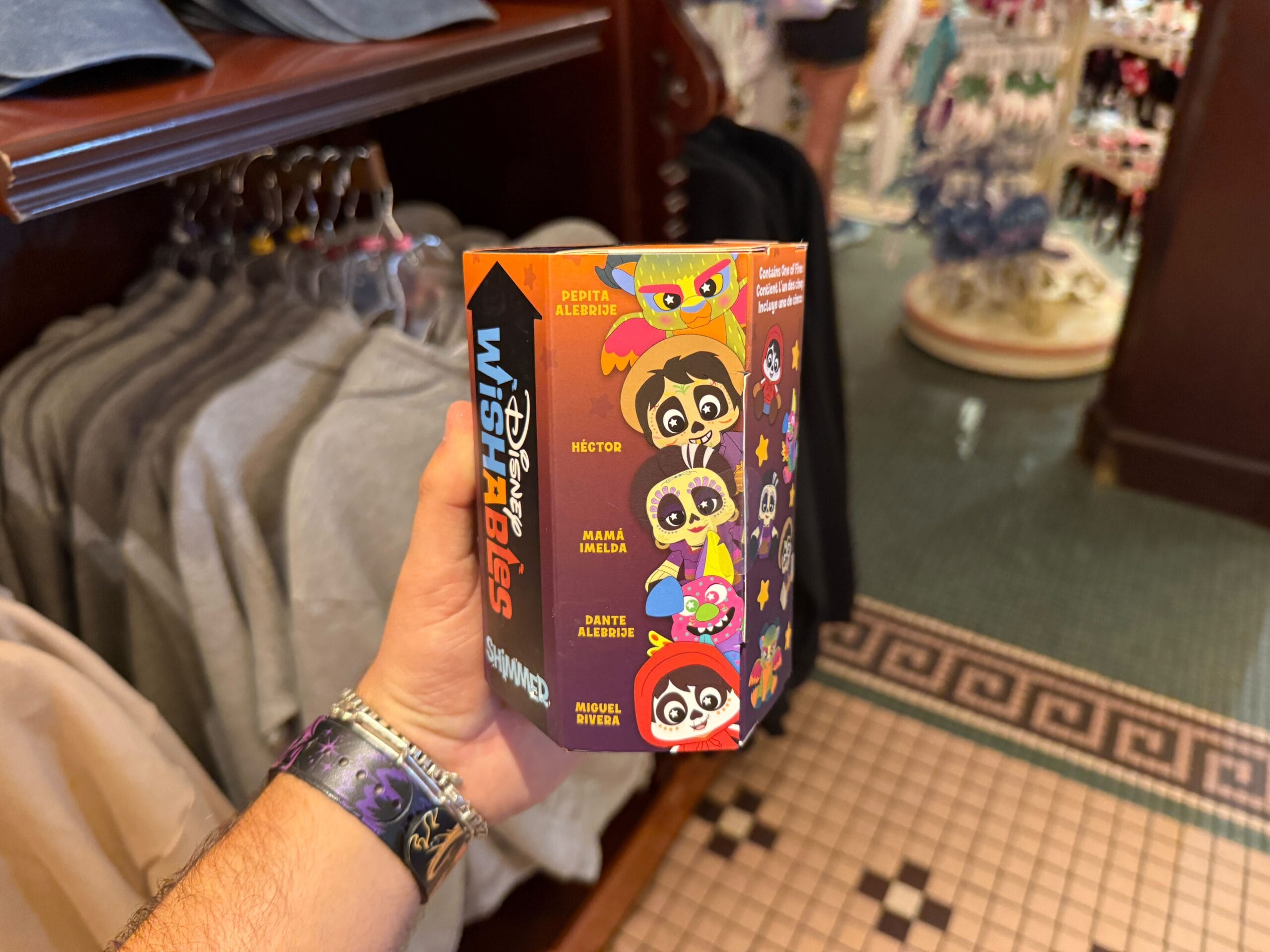 A hand holds a Coco Shimmer Wishables mystery plush box in a store at the Disneyland Resort near clothing racks.