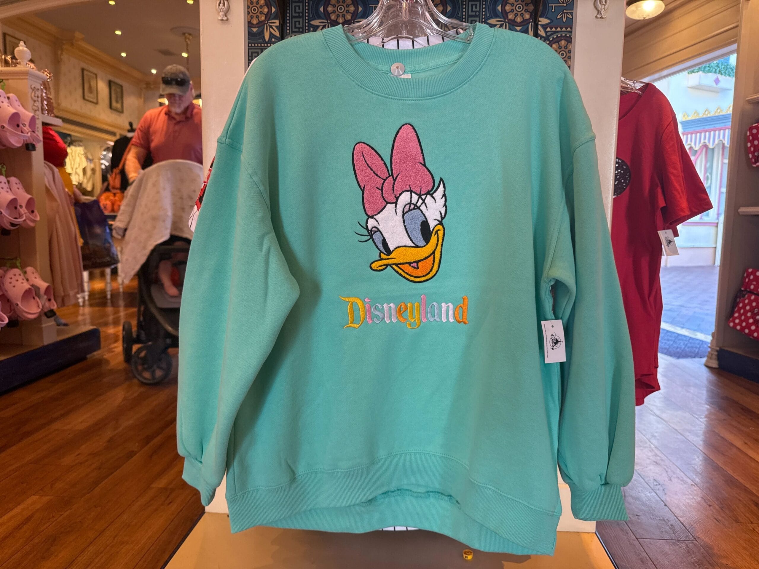 A green Disneyland Character Sweatshirt featuring Daisy Duck with a pink bow, displayed in a store.