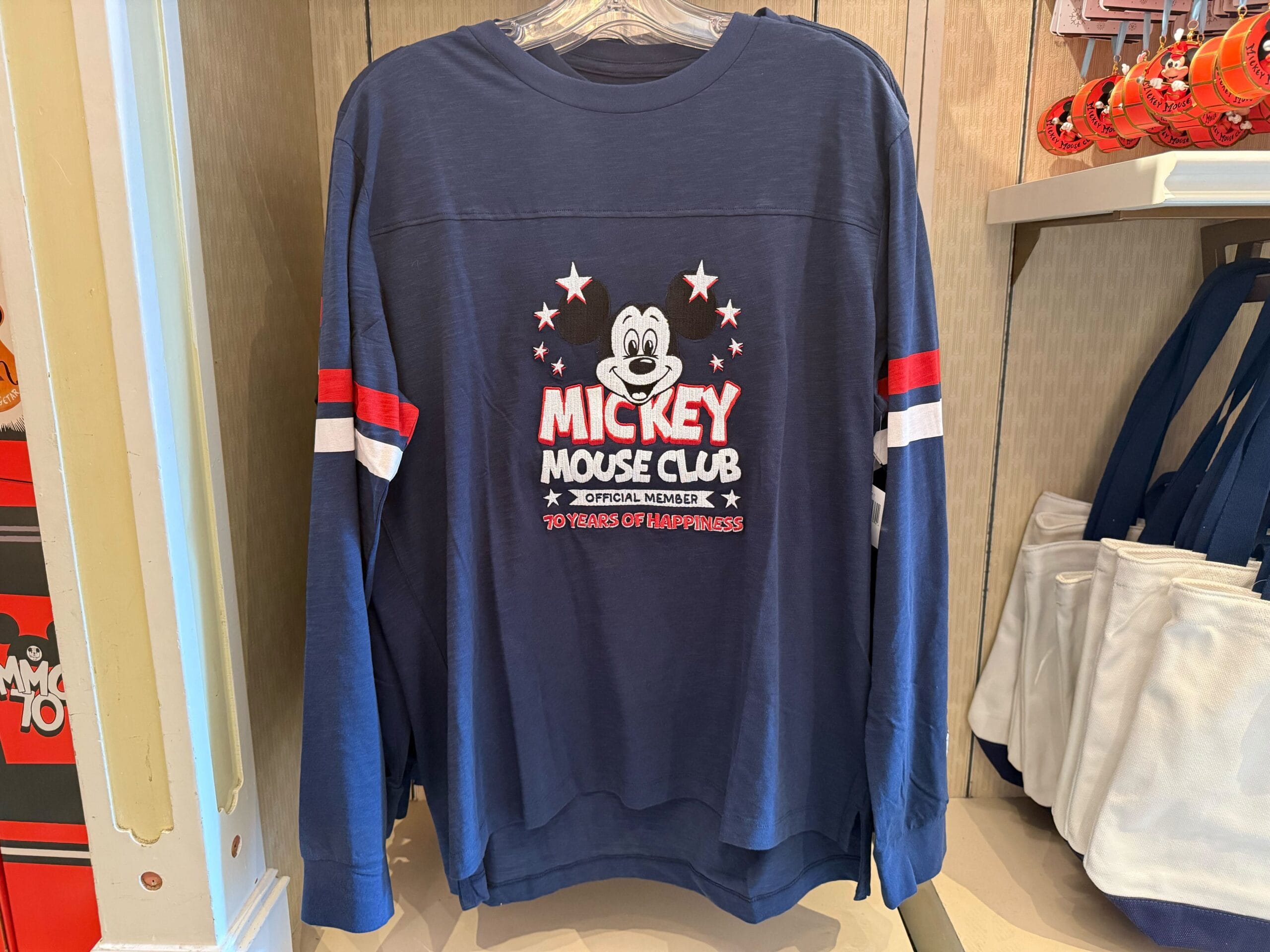 Blue long-sleeve shirt with Mickey Mouse Club merchandise logo and "70th anniversary" on display in a Disneyland store.