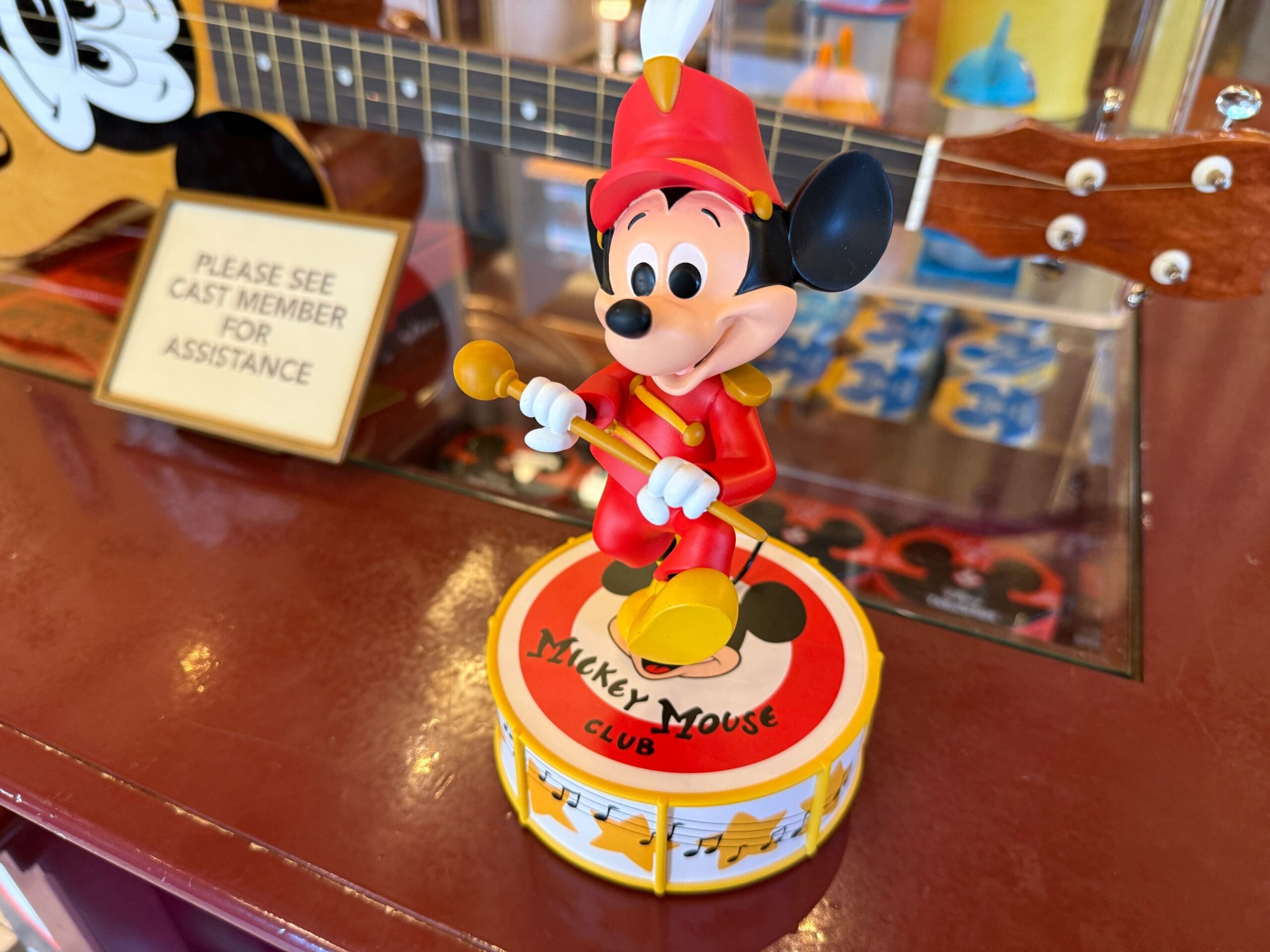 A Disneyland Mickey Mouse Club figurine in a red band outfit stands by a “Please See Cast Member” sign.