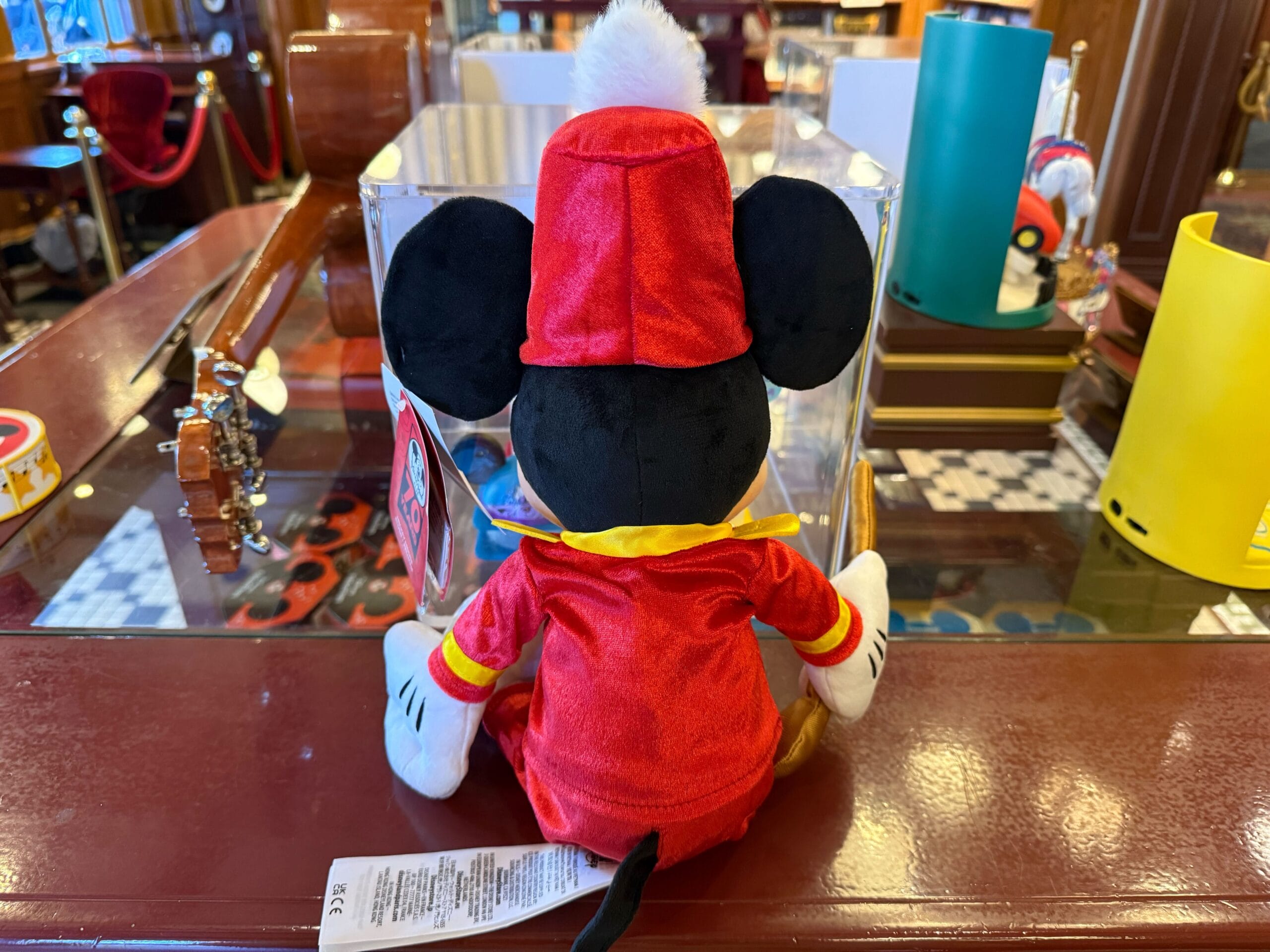 Plush Mickey Mouse in a red costume sits on a counter, surrounded by Disneyland 70th anniversary merchandise.