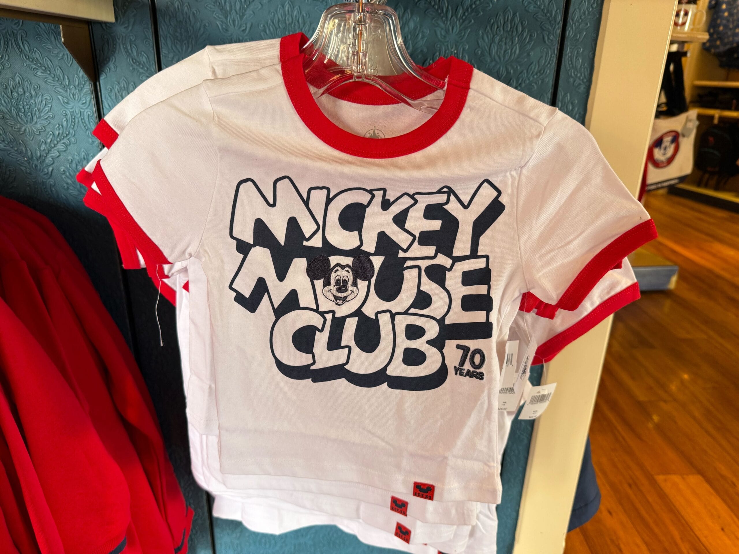White and red Disneyland T-shirt with "Mickey Mouse Club 70 Years" and Mickey's face, hanging on a rack.