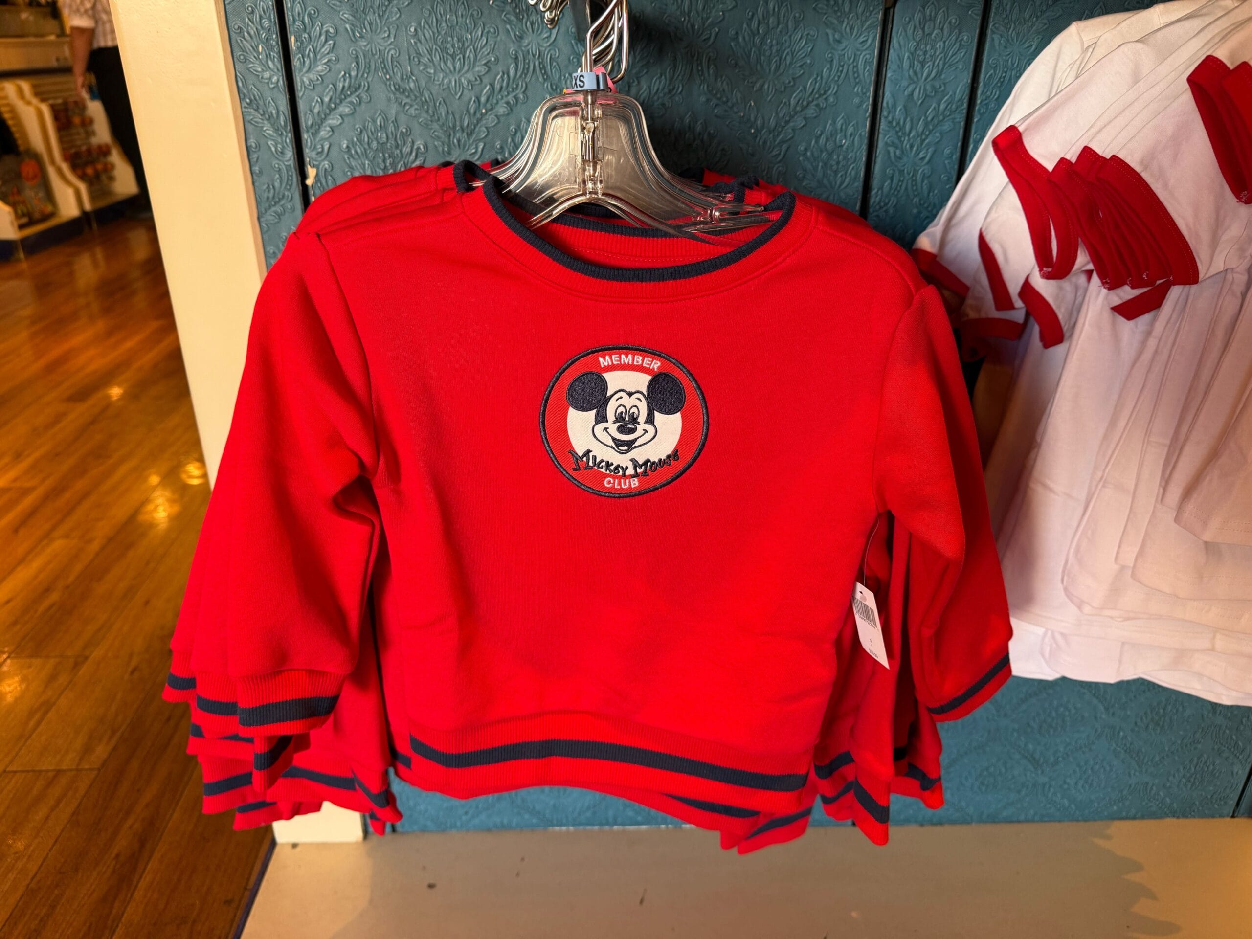 Red children's shirt with a Mickey Mouse patch celebrates the Mickey Mouse Club 70th Anniversary, hanging on a display rack.