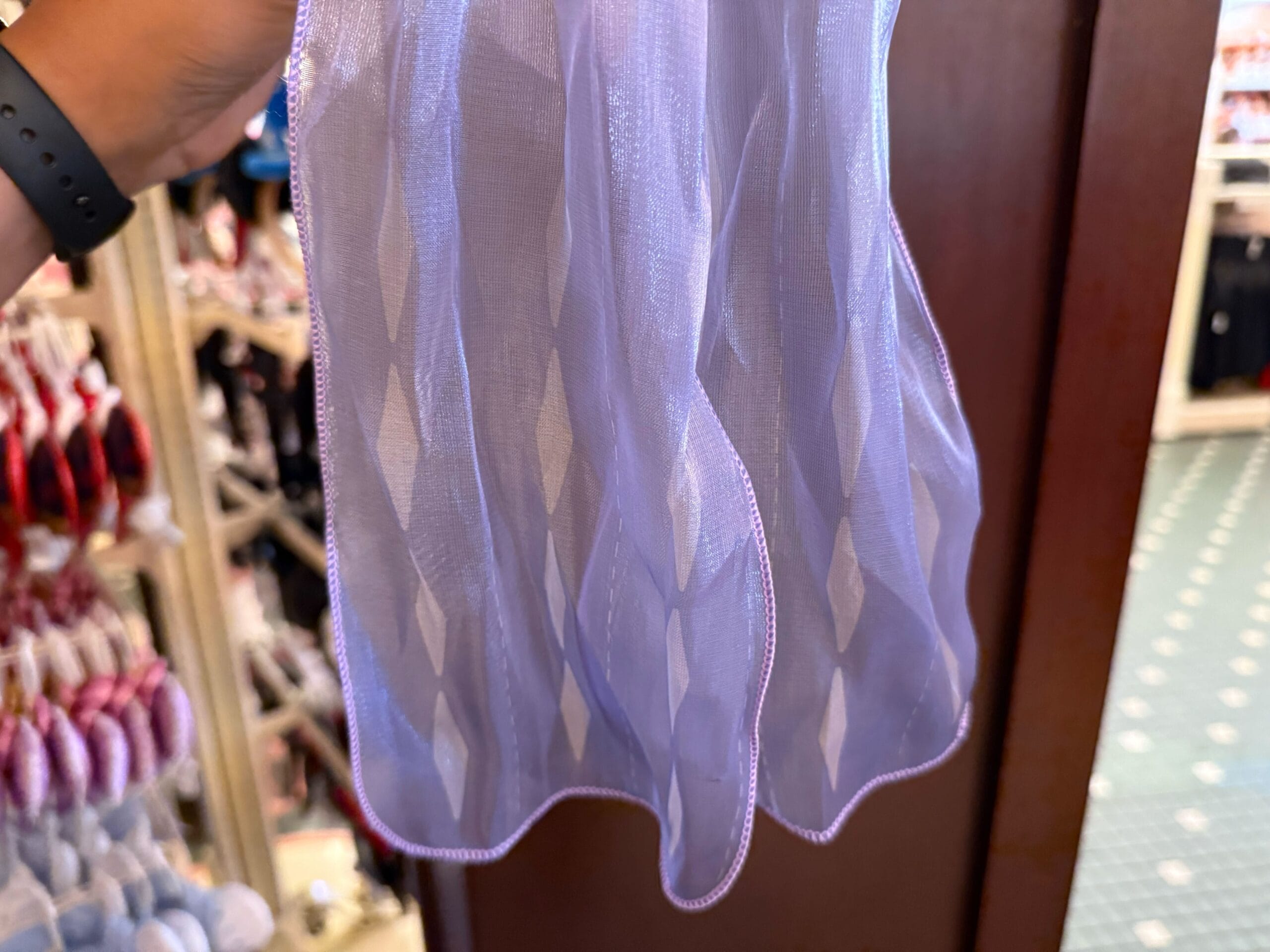 A hand holds sheer, light purple fabric with a diamond pattern, displayed in a Disneyland store with shelves in the background.