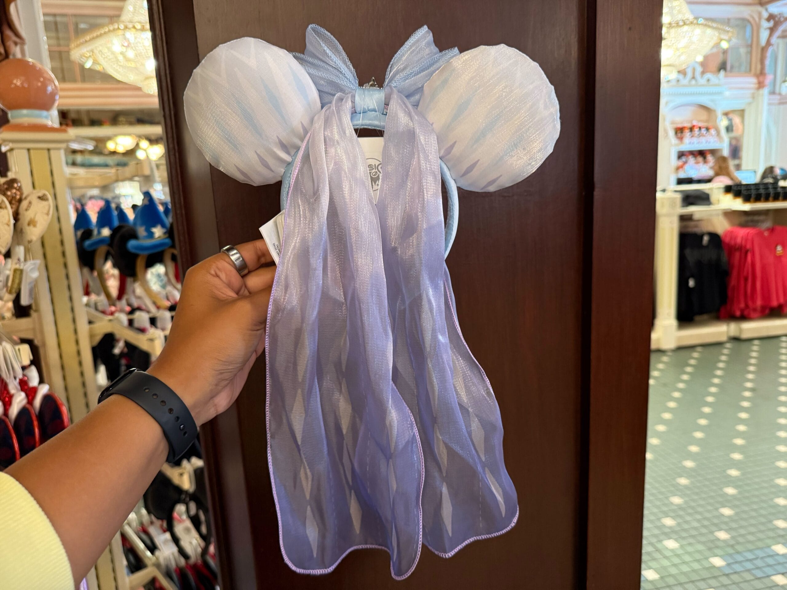 A hand holds a pastel blue Minnie Mouse ear headband with a long sheer veil in a Disneyland shop.