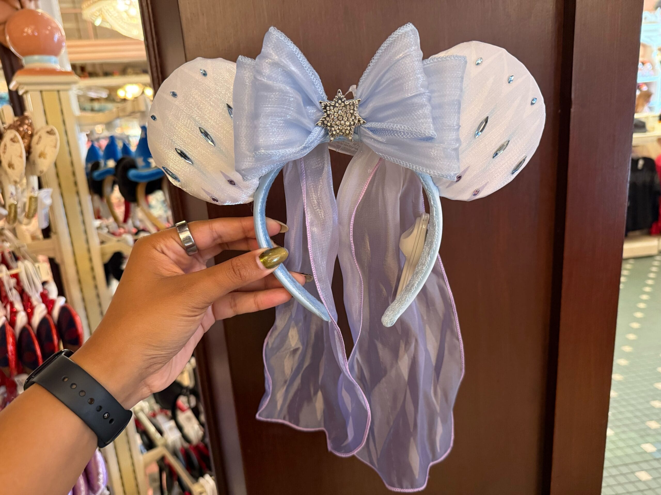 A hand holds a sparkly, light blue Disneyland Elsa Ear Headband with mouse ears, bow, and flowing sheer ribbons.
