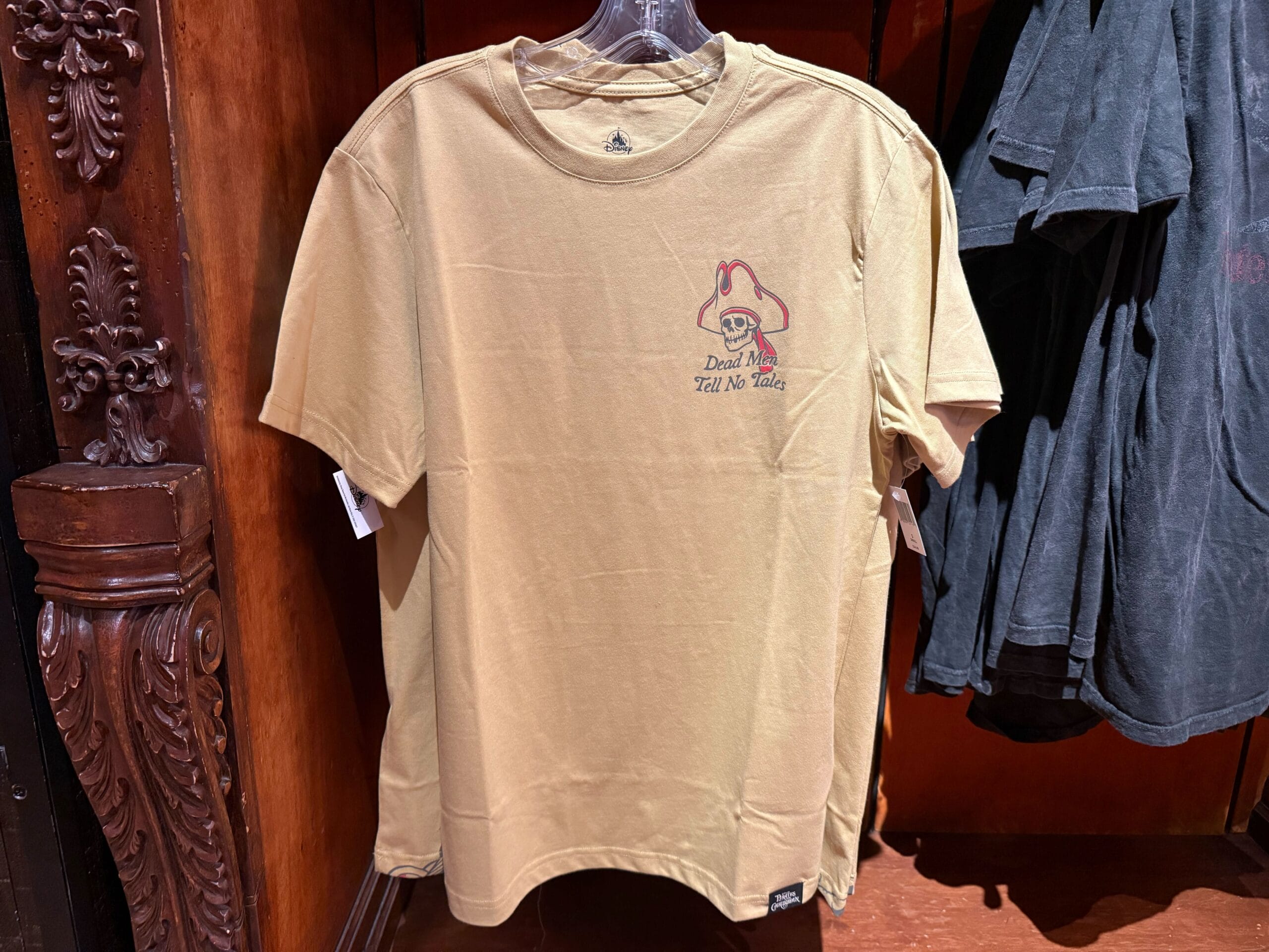 Beige t-shirt with a Pirates of the Caribbean skull graphic and "Dead Men Tell No Tales" text, hanging on display.