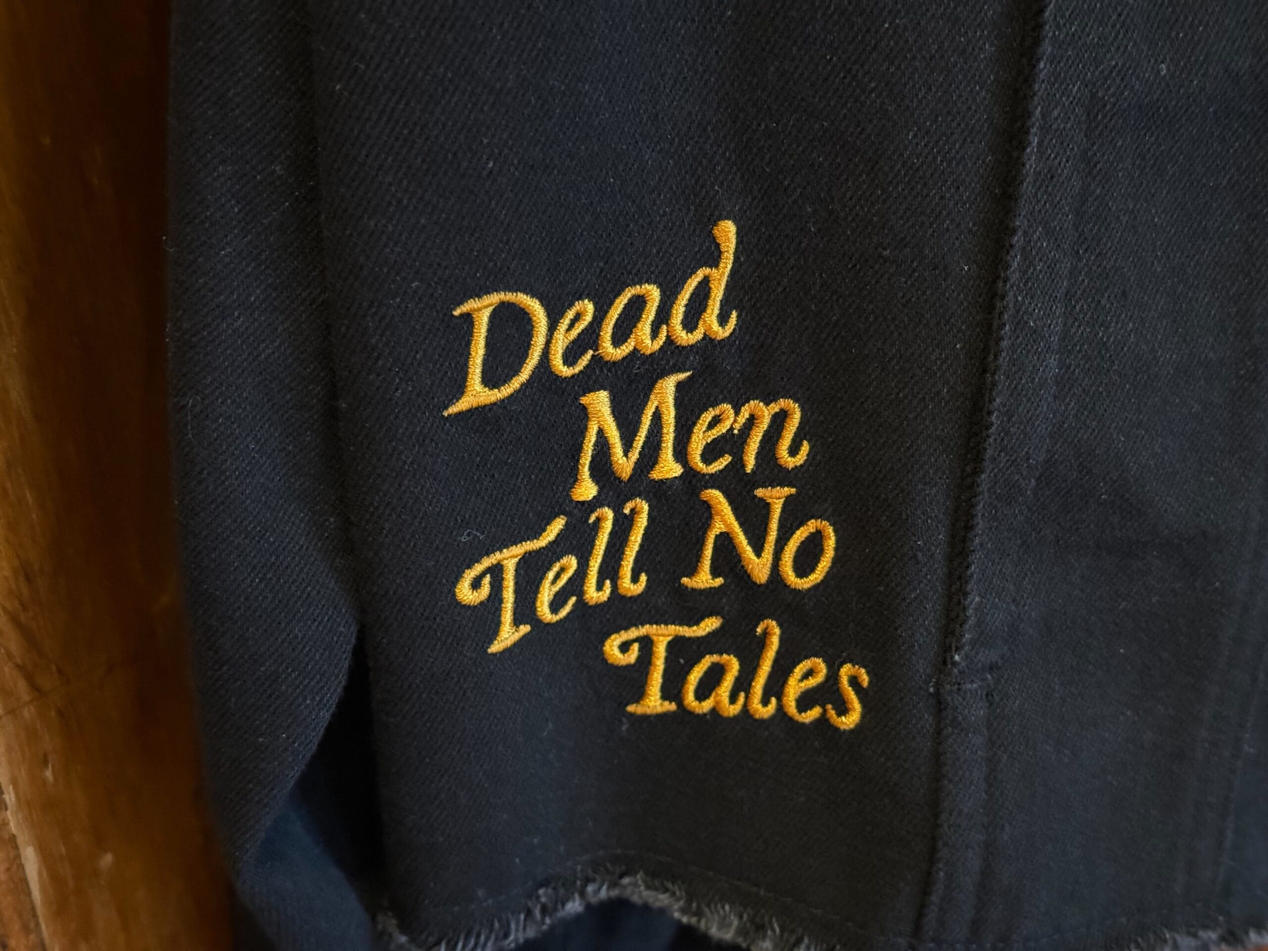Black fabric with yellow embroidered text reading, "Dead Men Tell No Tales"—perfect Pirates of the Caribbean apparel.