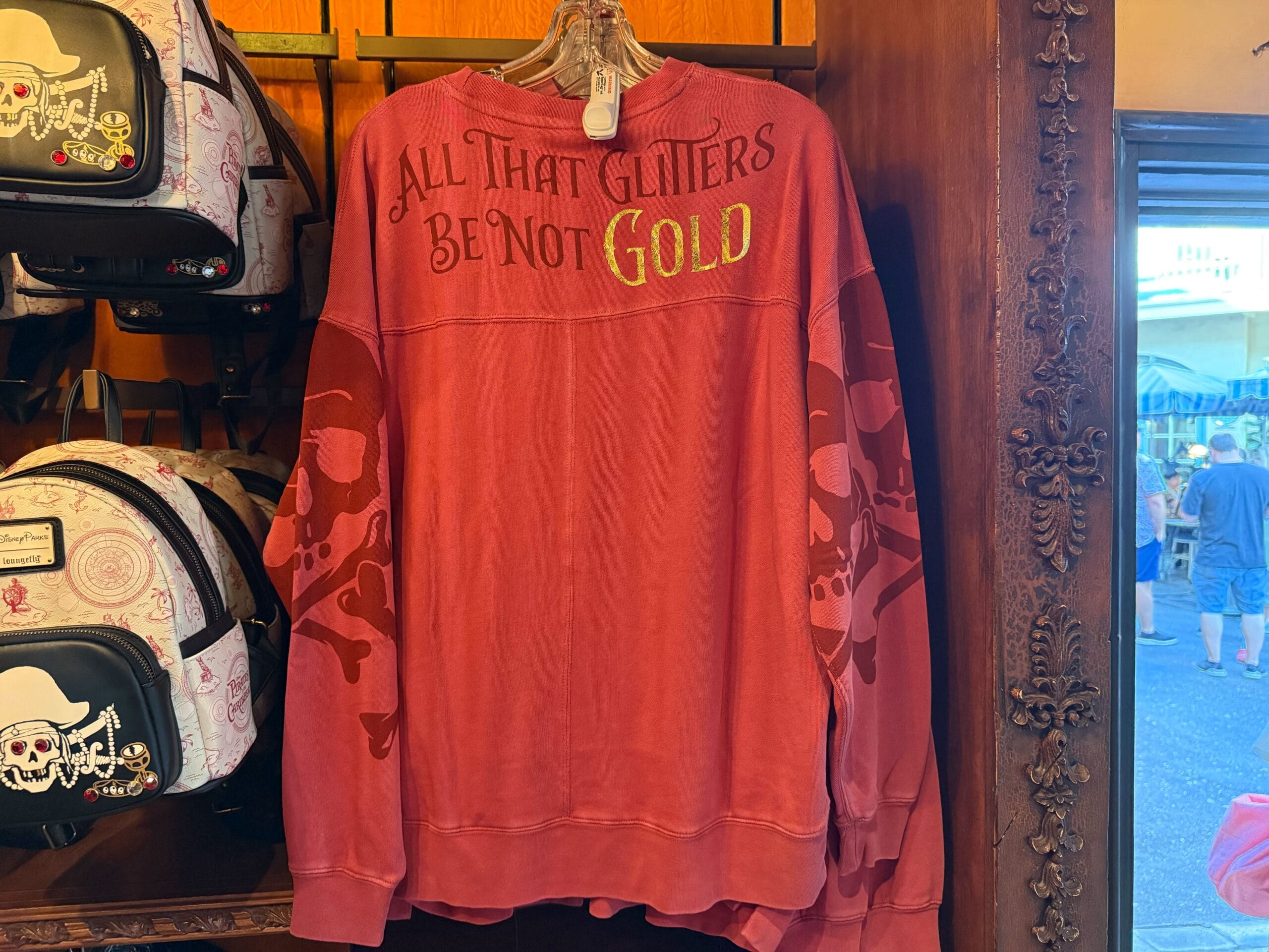 Red sweatshirt with gold and yellow text inspired by Pirates of the Caribbean, hanging on a store display rack.