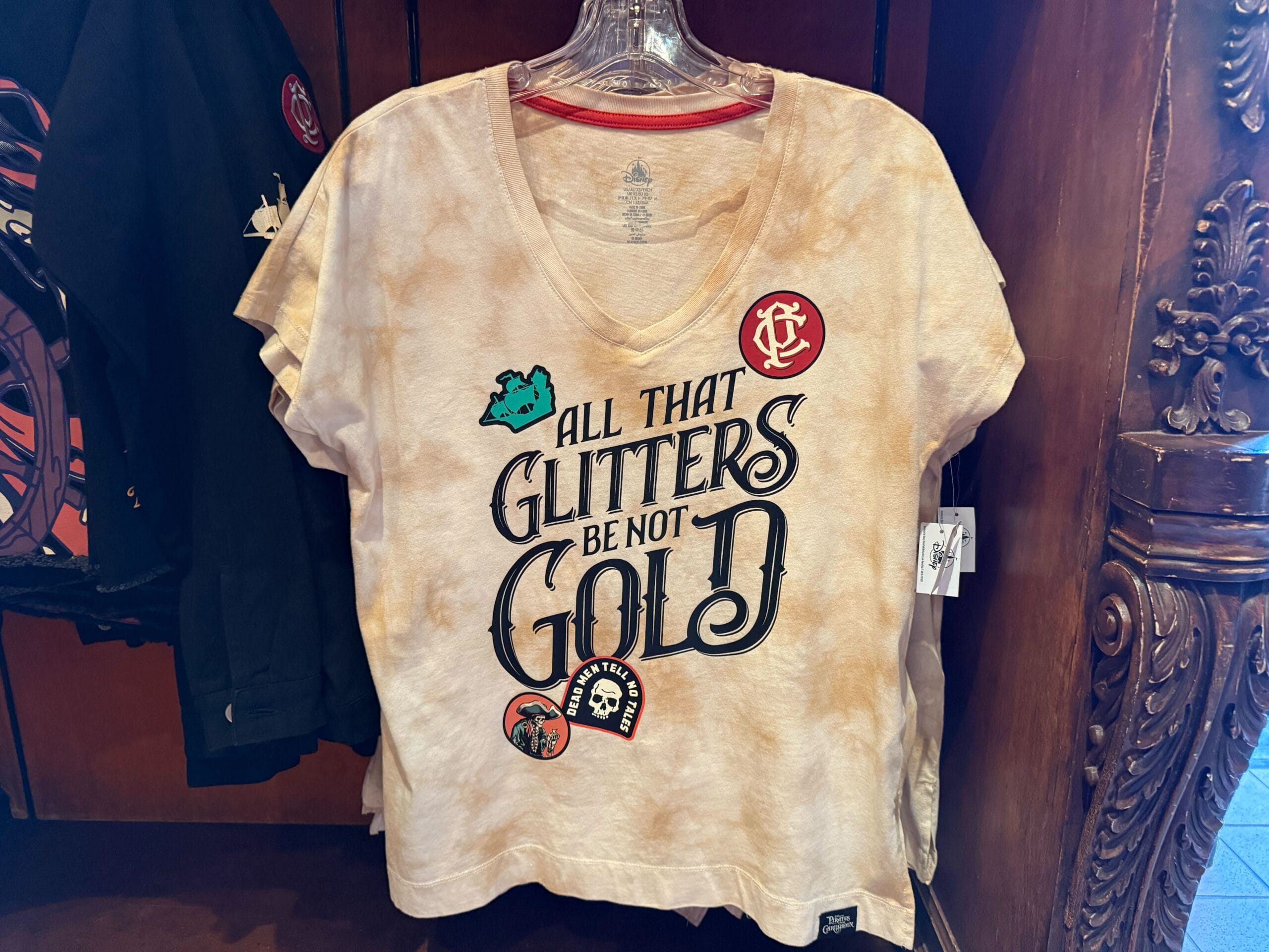 Tan t-shirt on hanger featuring “All that glitters be not gold” with pirate graphics—new Pirates of the Caribbean merchandise.