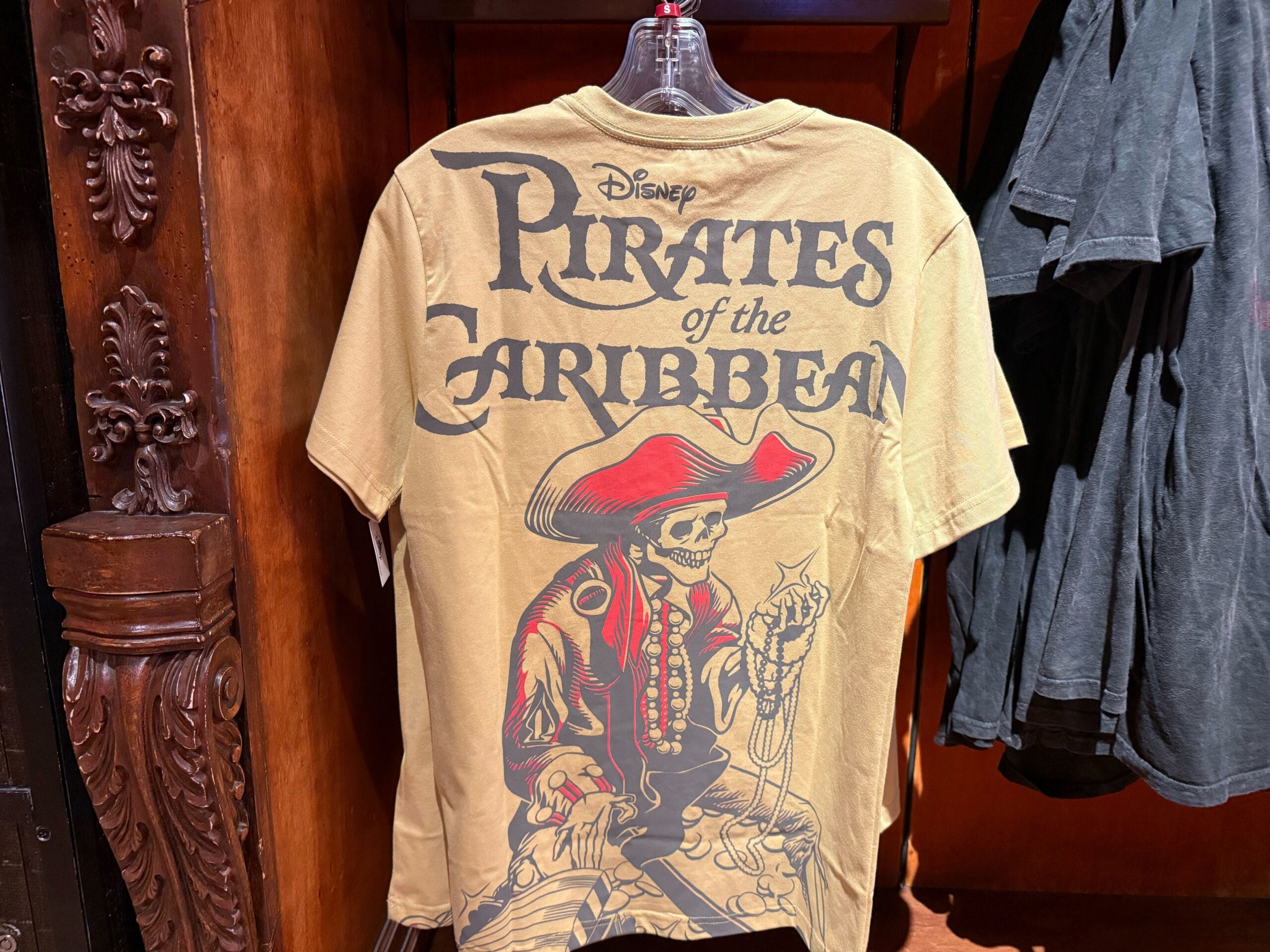 Tan t-shirt featuring a skeleton pirate and bold "Pirates of the Caribbean" text—perfect Disneyland apparel.