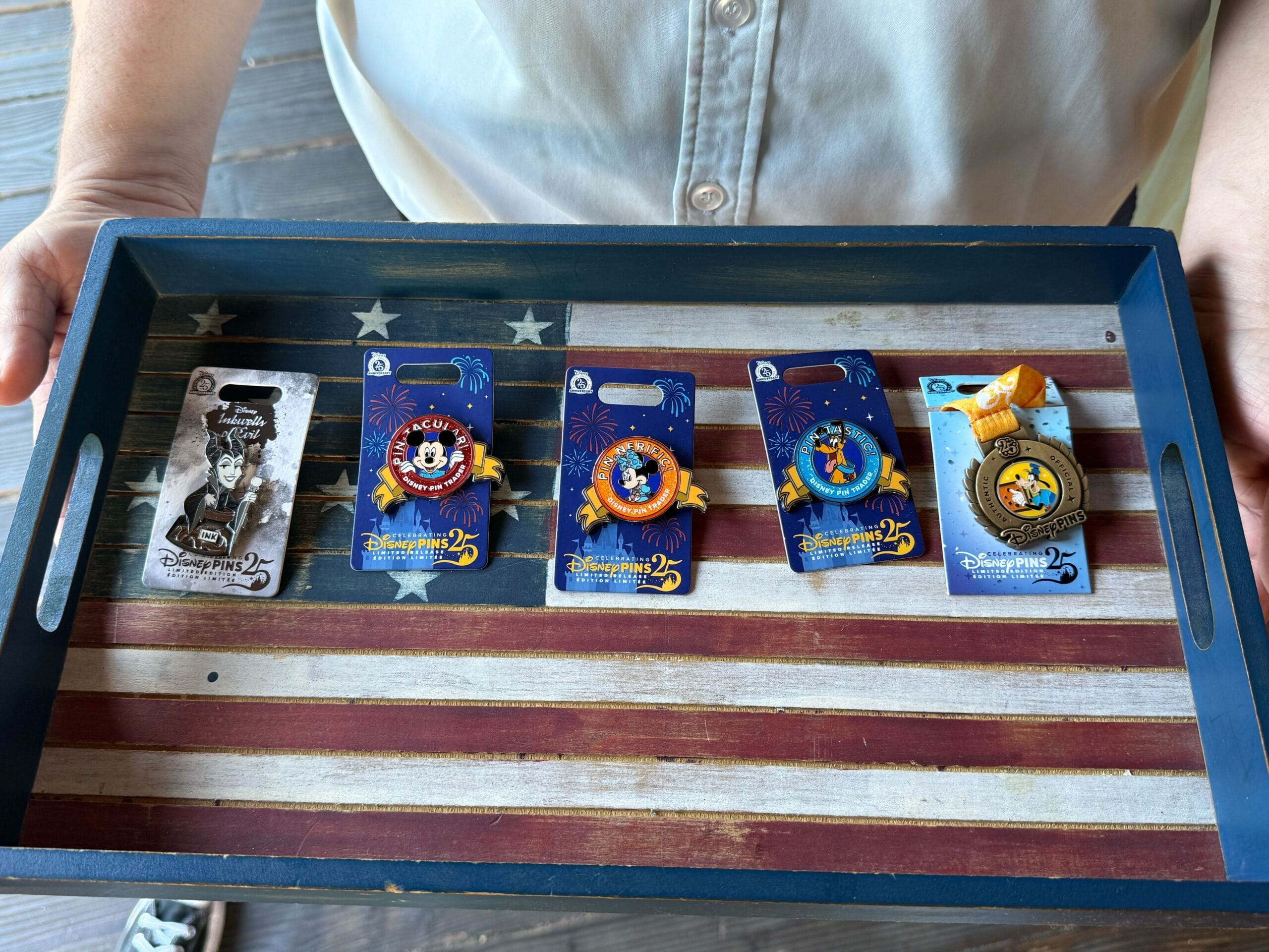 A person holds a tray with an American flag design, displaying five Disneyland Pin Trading 25th Anniversary pins.