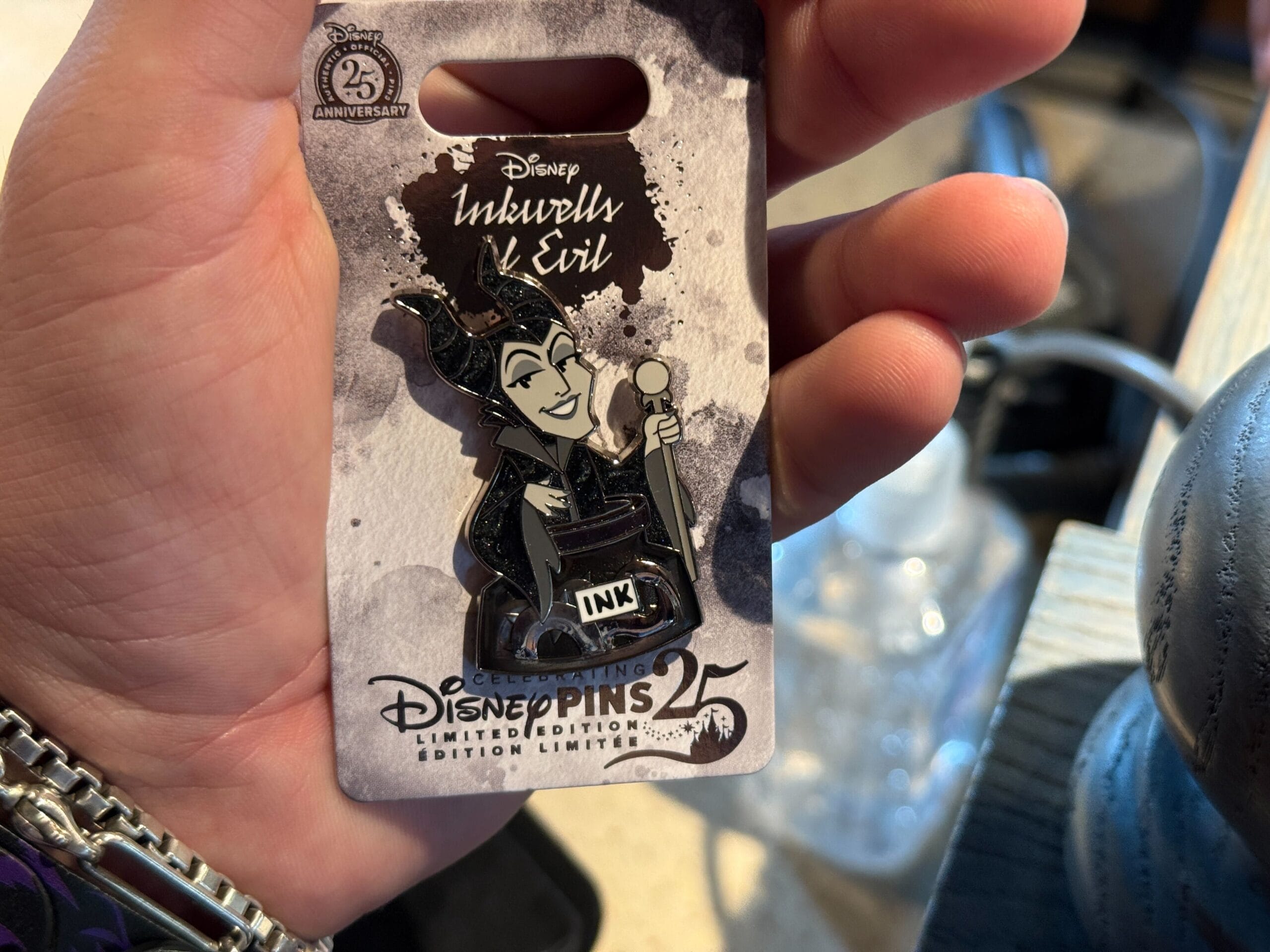 A hand holds a Disney pin featuring Maleficent, labeled "Inkwell & Evil," for Disneyland Pin Trading's 25th anniversary.