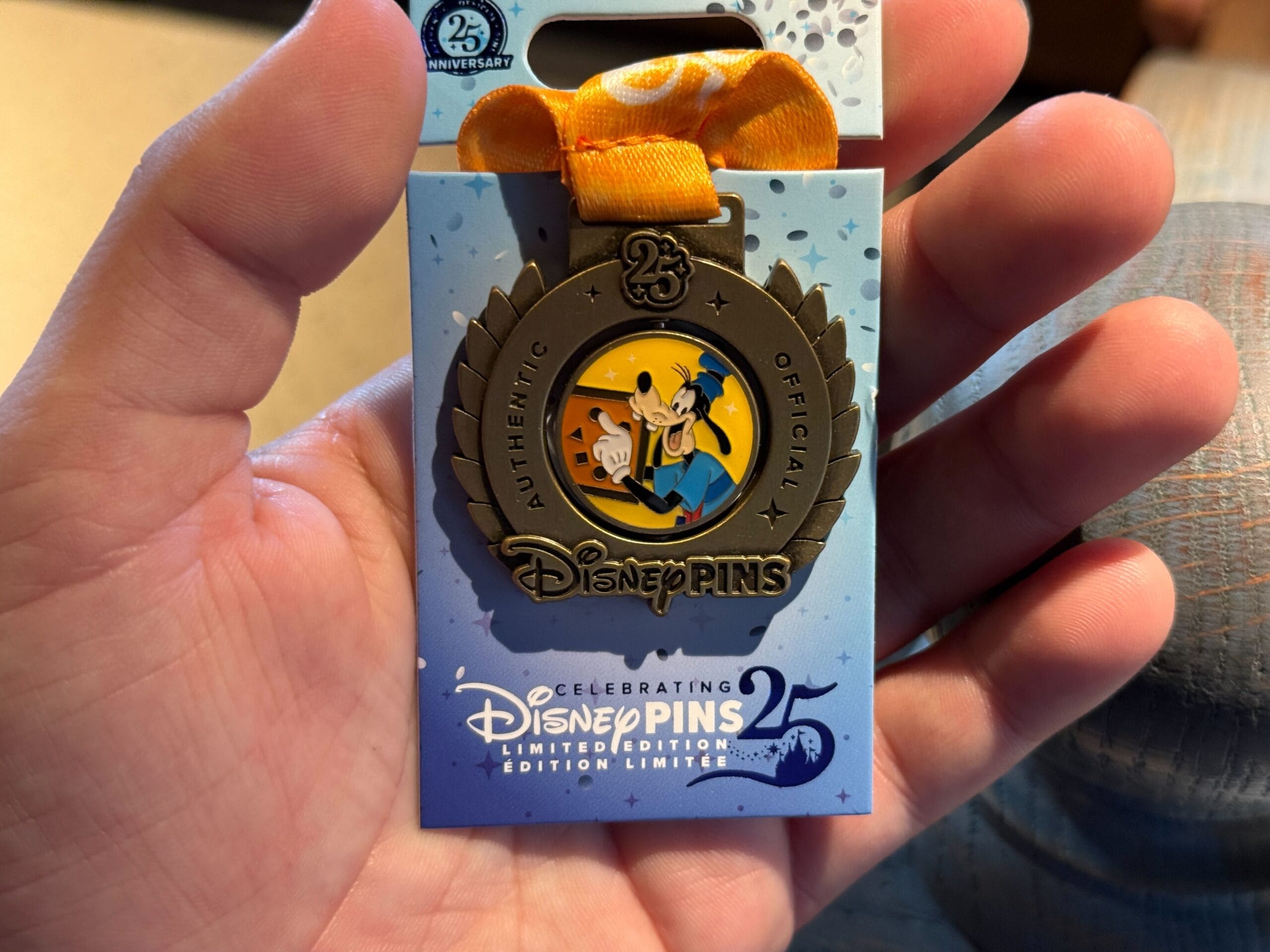 A hand holds a Pin Trading 25th Anniversary pin with Mickey and Friends on a blue card, adorned with an orange ribbon.
