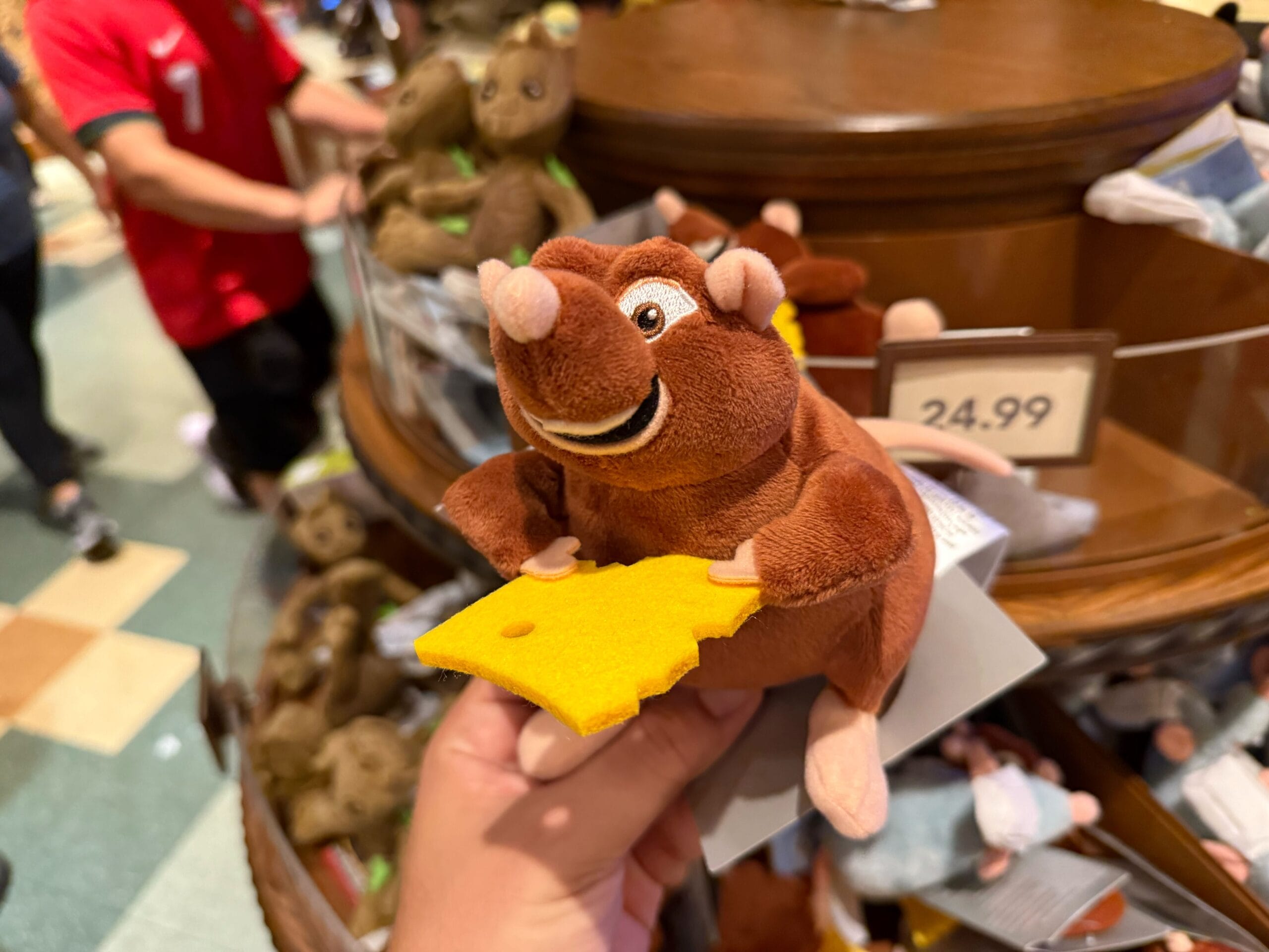 A hand holds an Emile Shoulder Plush rat toy with yellow cheese in front of a display of stuffed animals.
