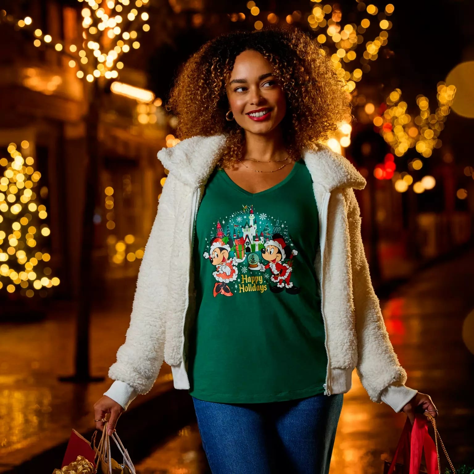 Woman wearing a festive “Happy Holidays” shirt, holding Disney Christmas Merchandise shopping bags under 2025 holiday lights.