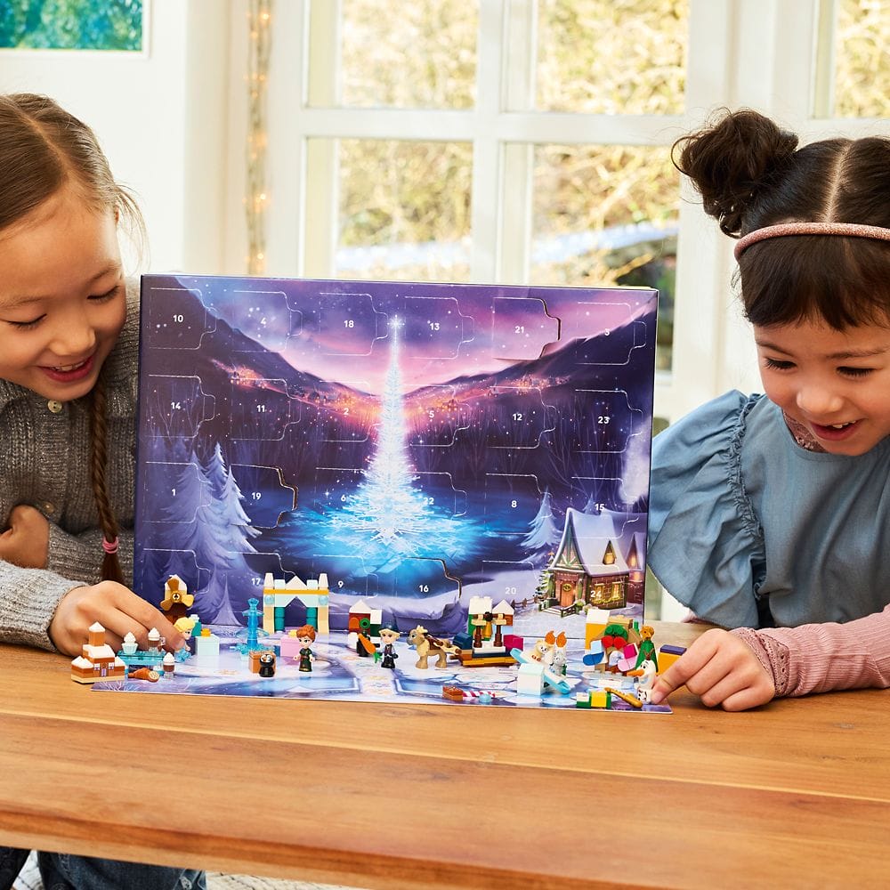 Two young girls play with LEGO toys in front of an advent calendar box with a snowy winter scene on it.