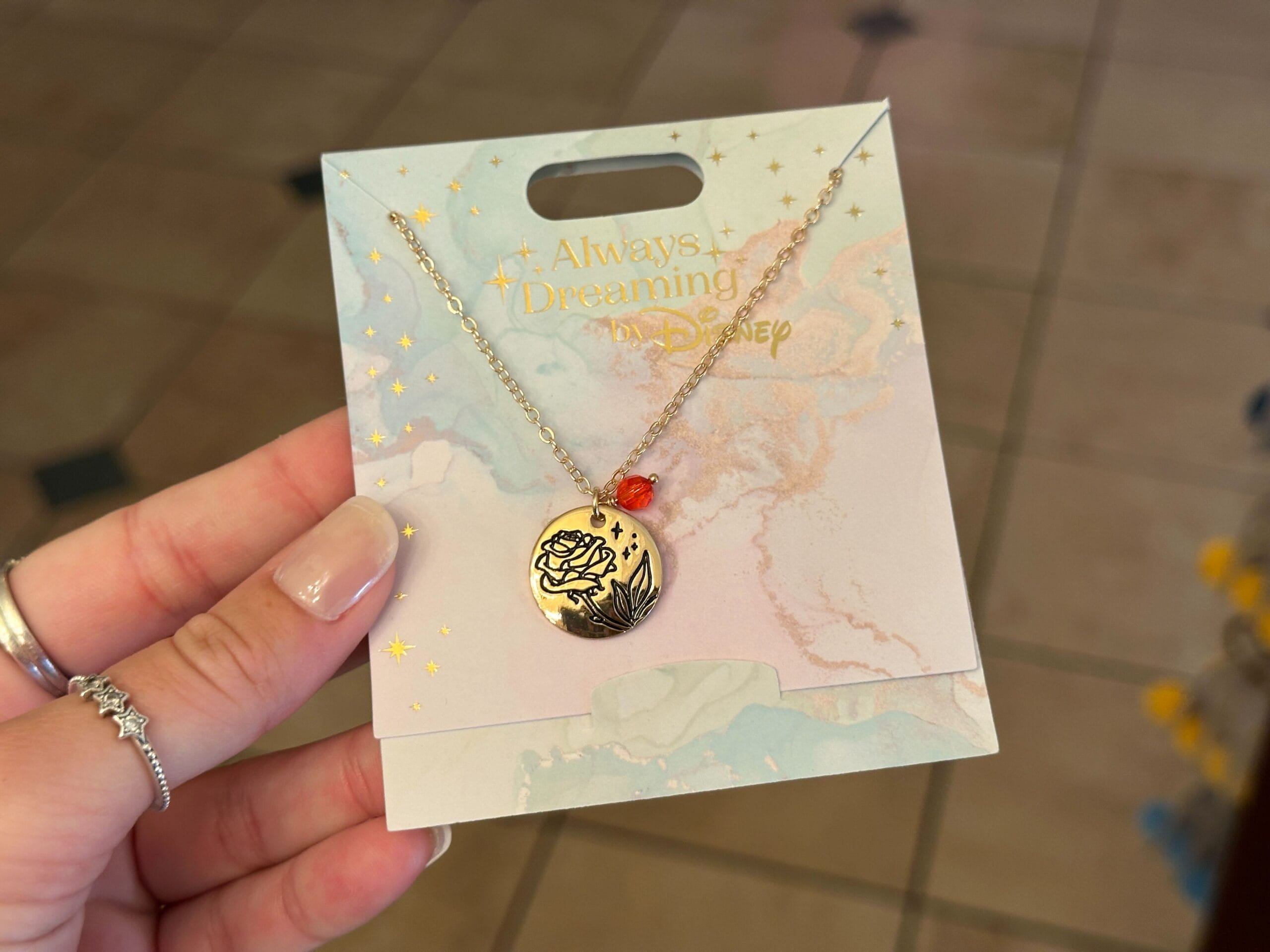 A hand holds a Disney jewelry collection necklace with a rose pendant and red bead on an "Always Dreaming" card.