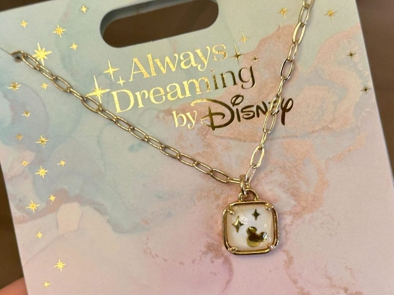 A hand holds a gold Tinker Bell pendant necklace from the Disney jewelry collection, on “Always Dreaming by Disney” packaging.