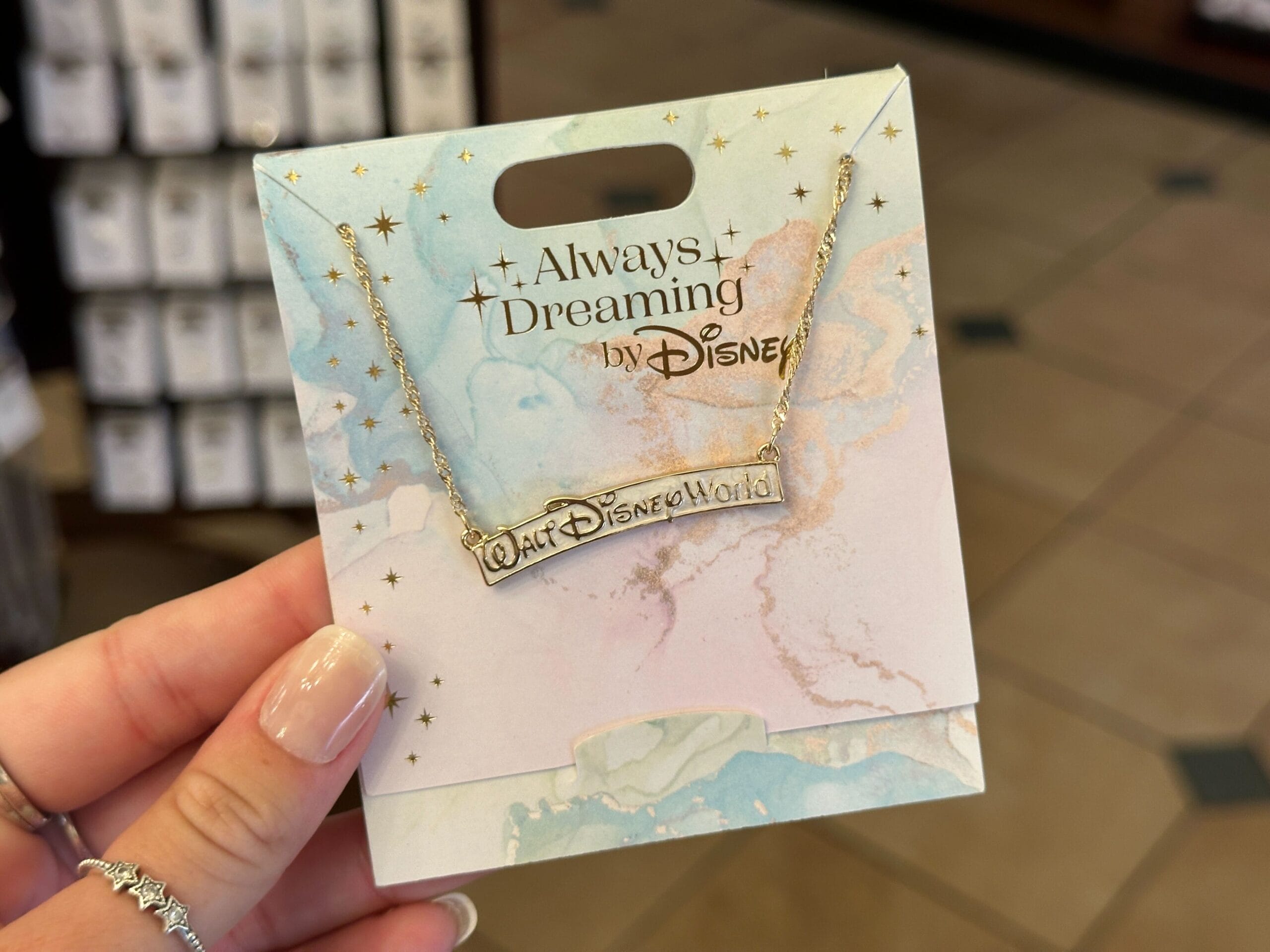 A hand holds a Disney necklace from the Disney jewelry collection on a pastel “Always Dreaming” card.