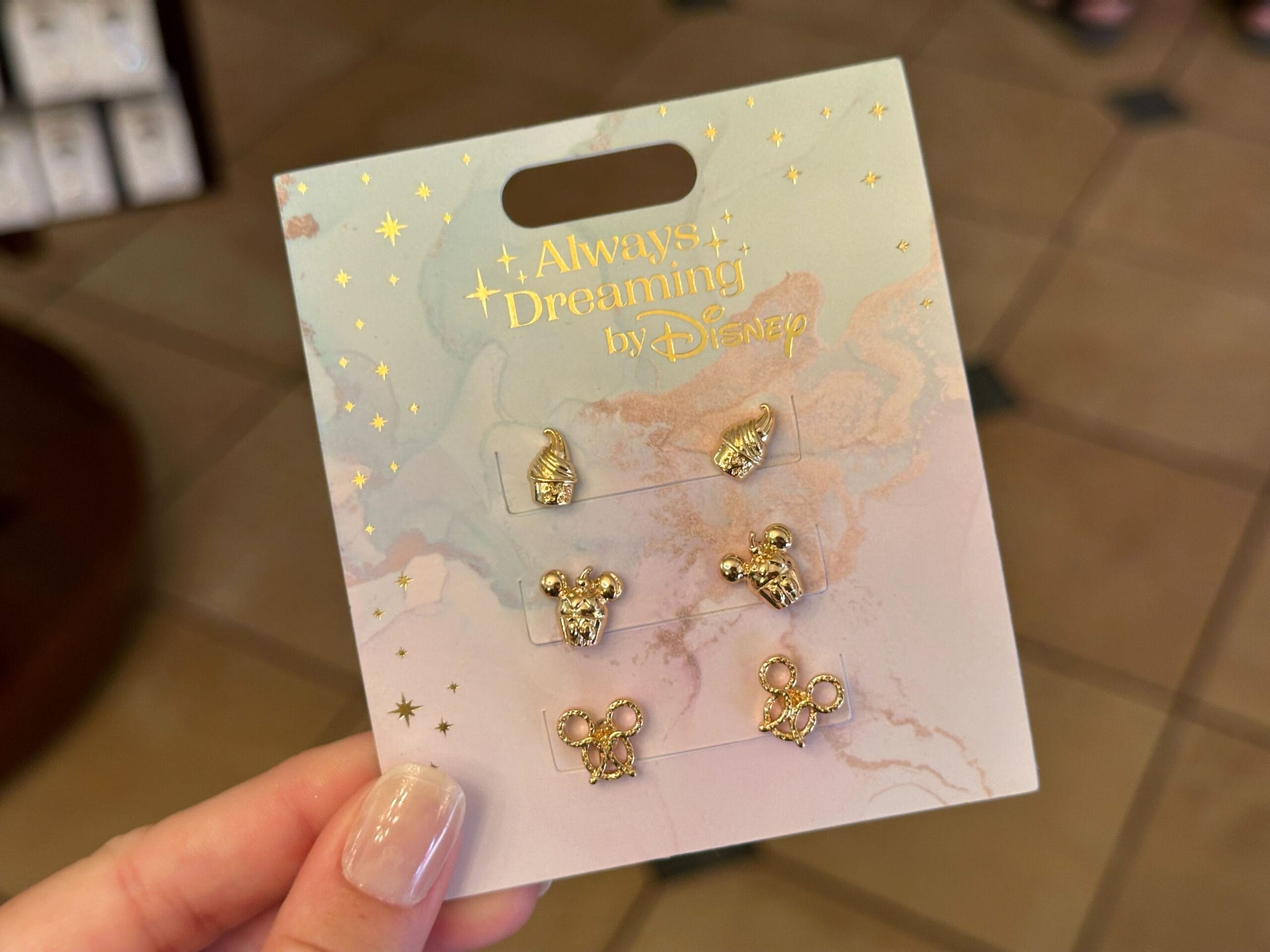 A hand holds a Disney accessories earring set with gold Mickey and Minnie on a pastel "Always Dreaming" card.
