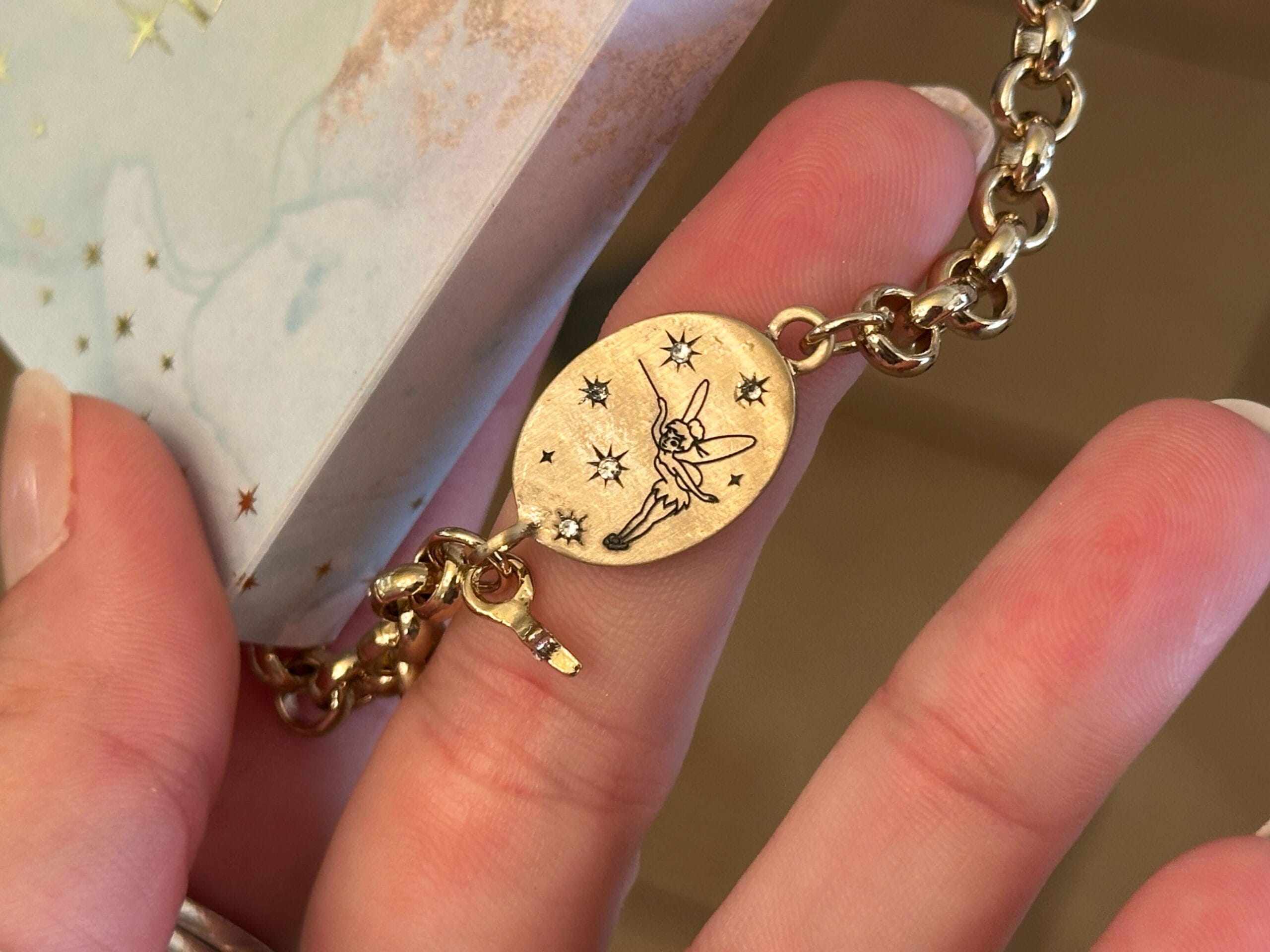 A gold bracelet charm from the Disney jewelry collection, engraved with a fairy and stars, held between manicured fingers.