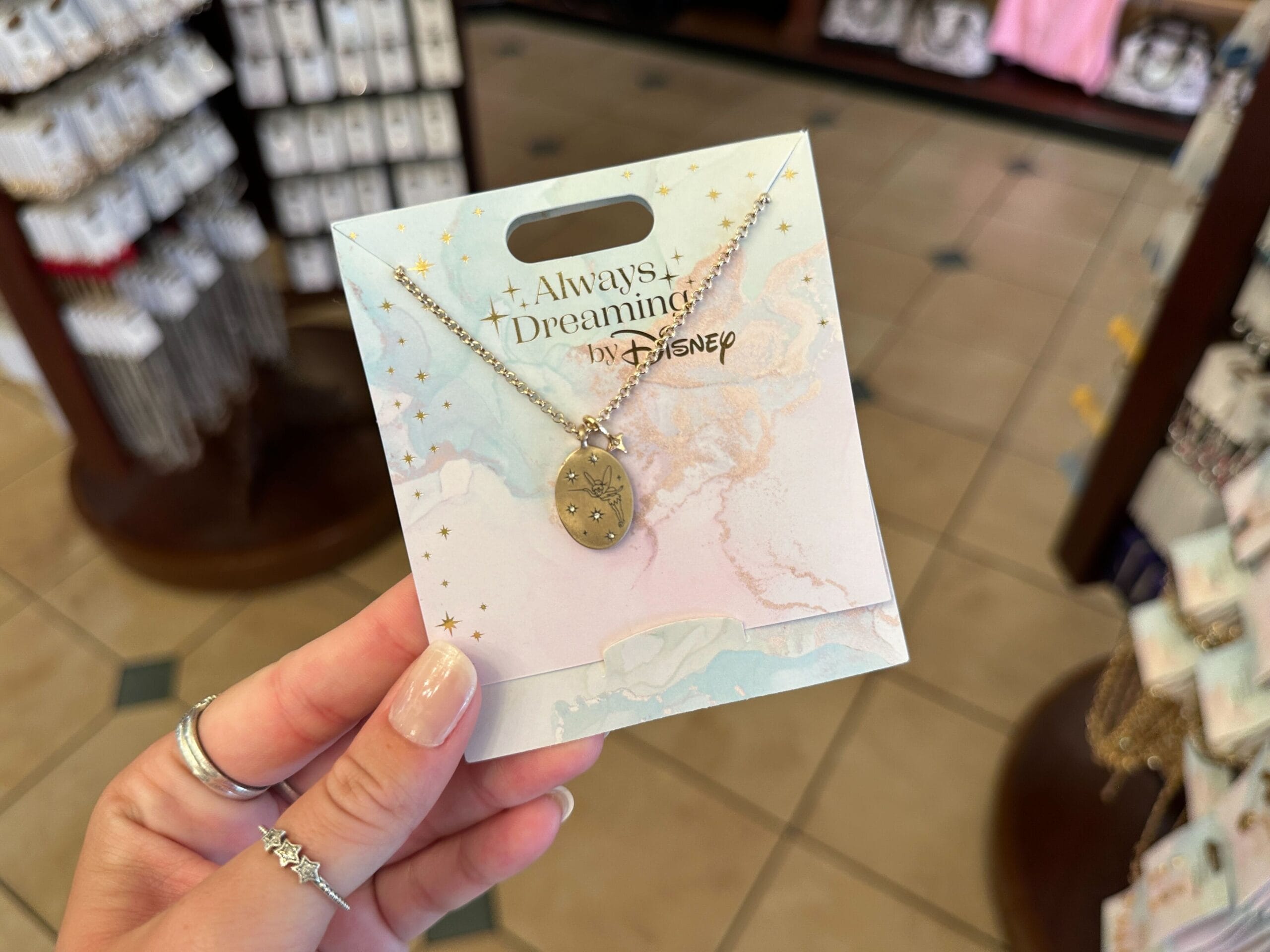 A hand holds a Disney “Always Dreaming” necklace from the Disney jewelry collection in a store with jewelry displays.