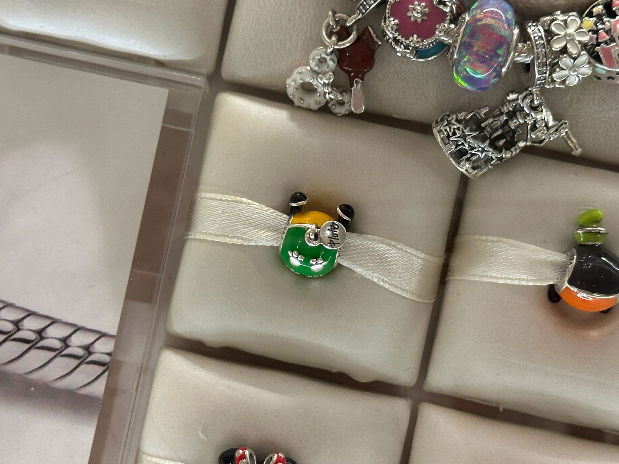A green, yellow, and black ladybug Pandora Charm with a silver tag, displayed on cream ribbon among other charms.