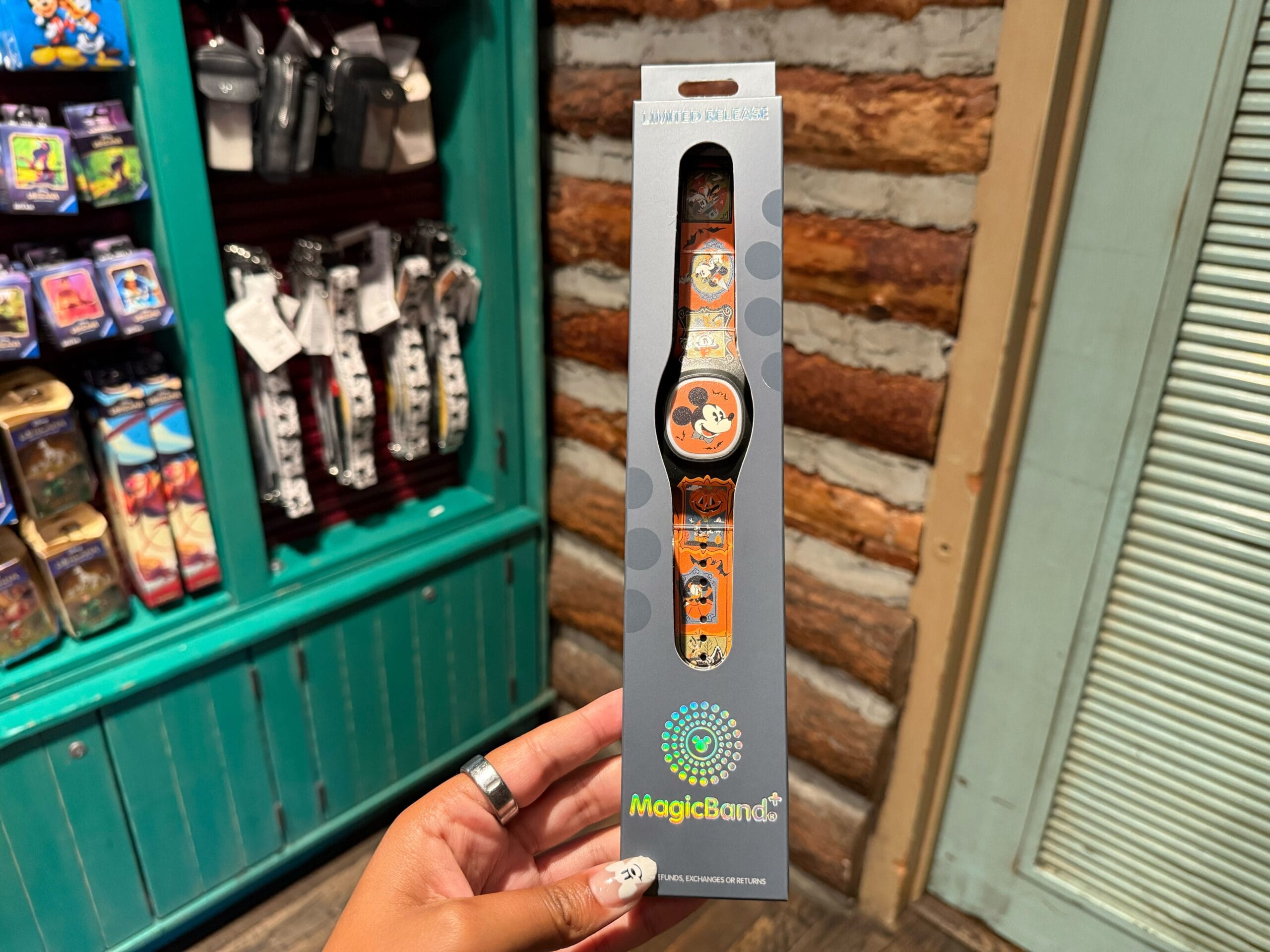 A hand holds a Disneyland Halloween MagicBand+ with Mickey Mouse designs, surrounded by shelves of themed merchandise.