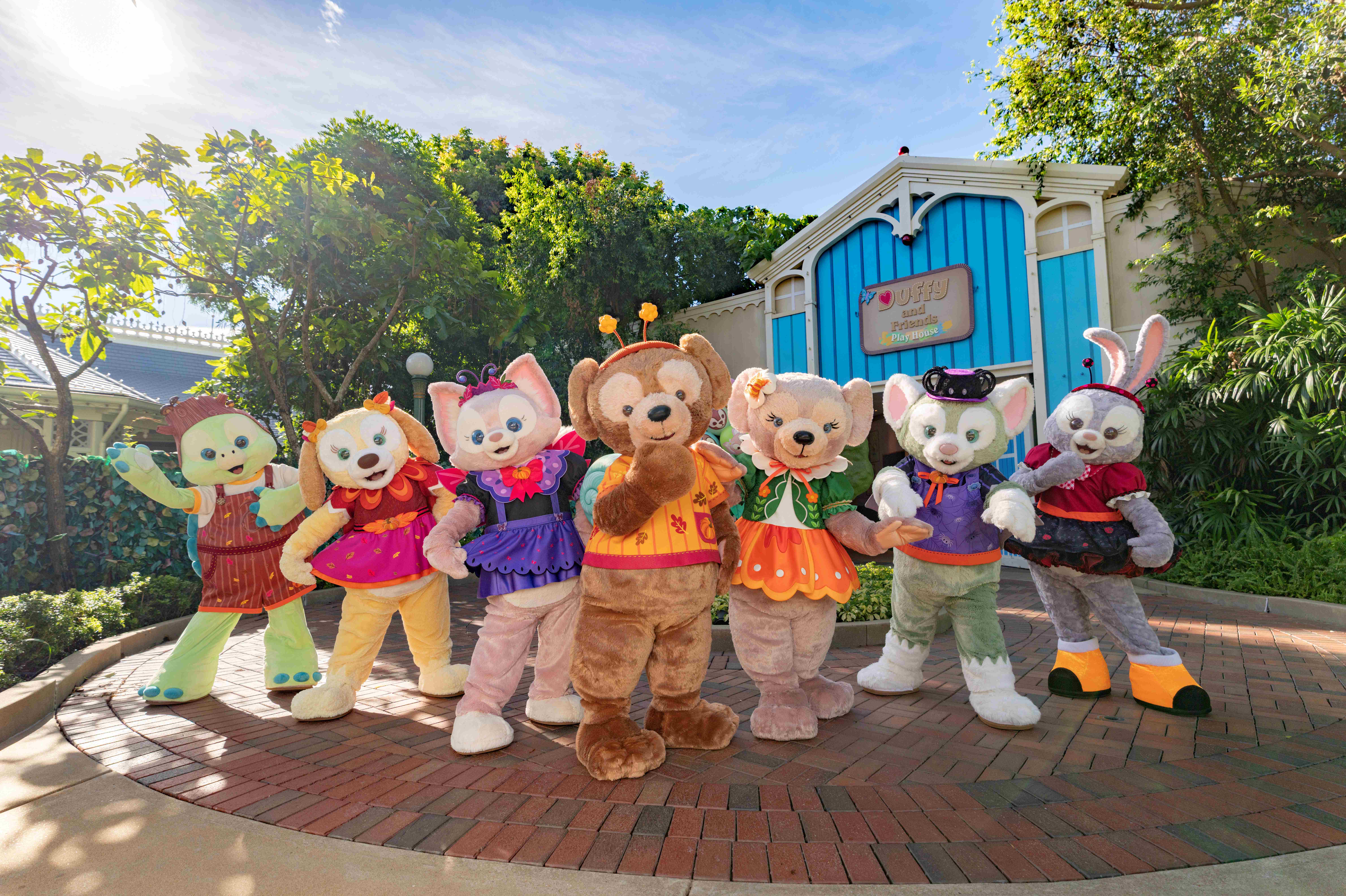 Seven colorful Disney Villains in costumes pose outside Hong Kong Disneyland on a sunny day near a blue building.