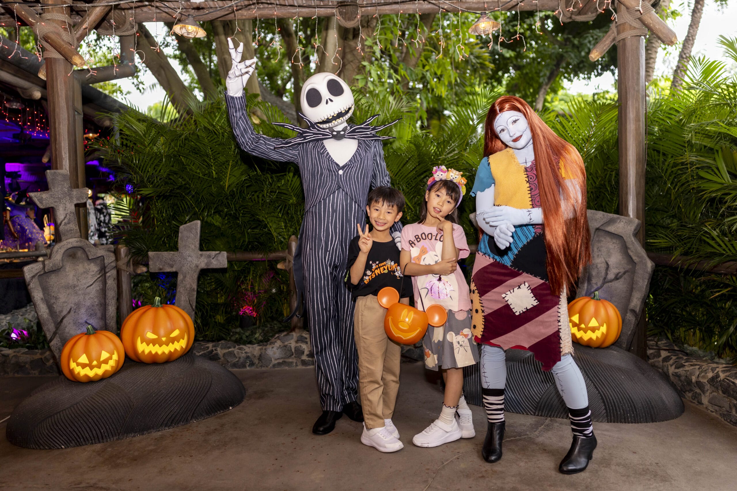 Two kids pose with Jack Skellington and Sally at Hong Kong Disneyland, surrounded by pumpkins and gravestones for Halloween.