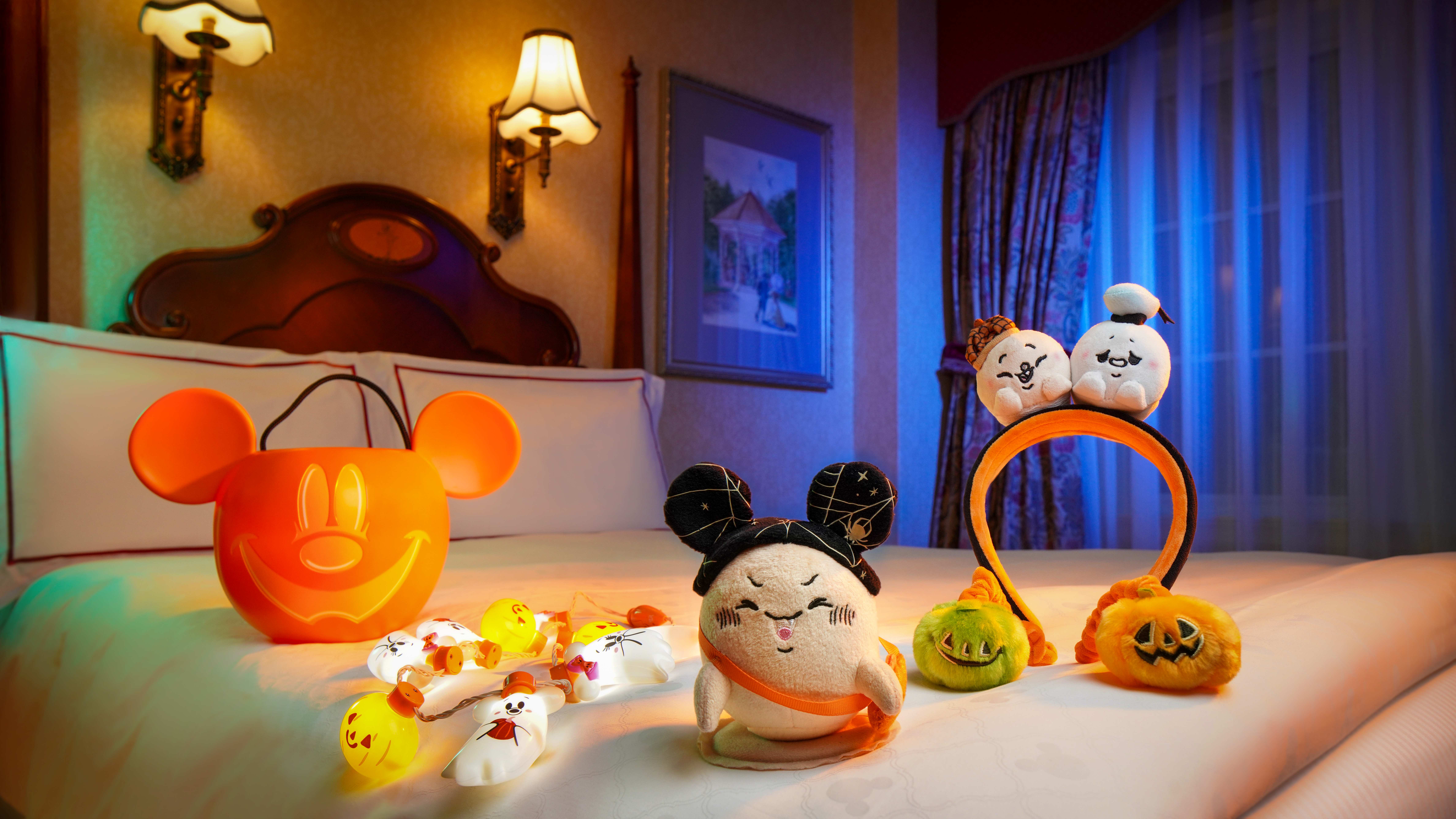 Halloween-themed Disney plush toys, glowing ghost figurines, and a Mickey pumpkin bucket from Hong Kong Disneyland on a hotel bed.
