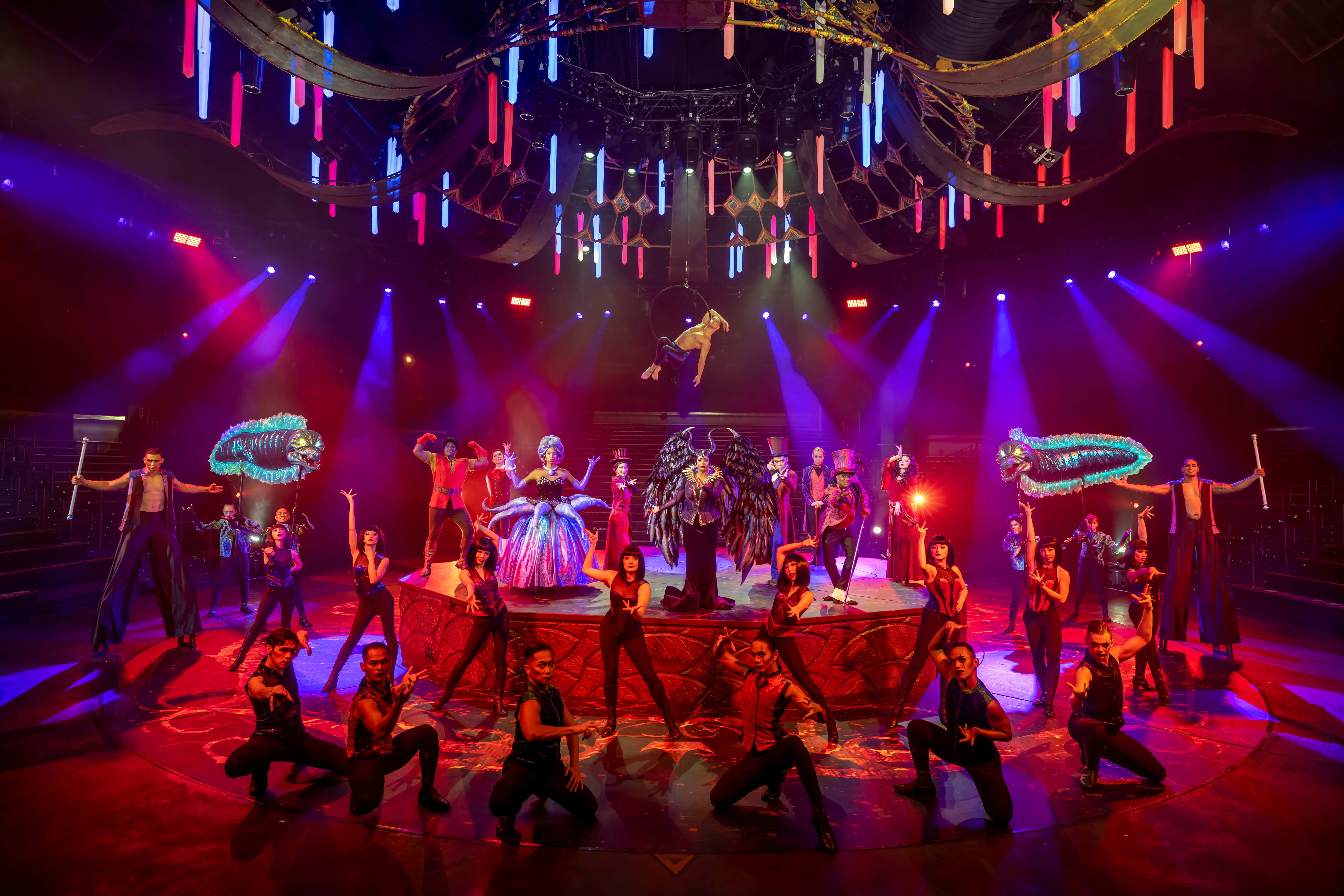 Circus performers pose dramatically under colorful lights, with Disney Villains-inspired costumes in a vibrant setting.