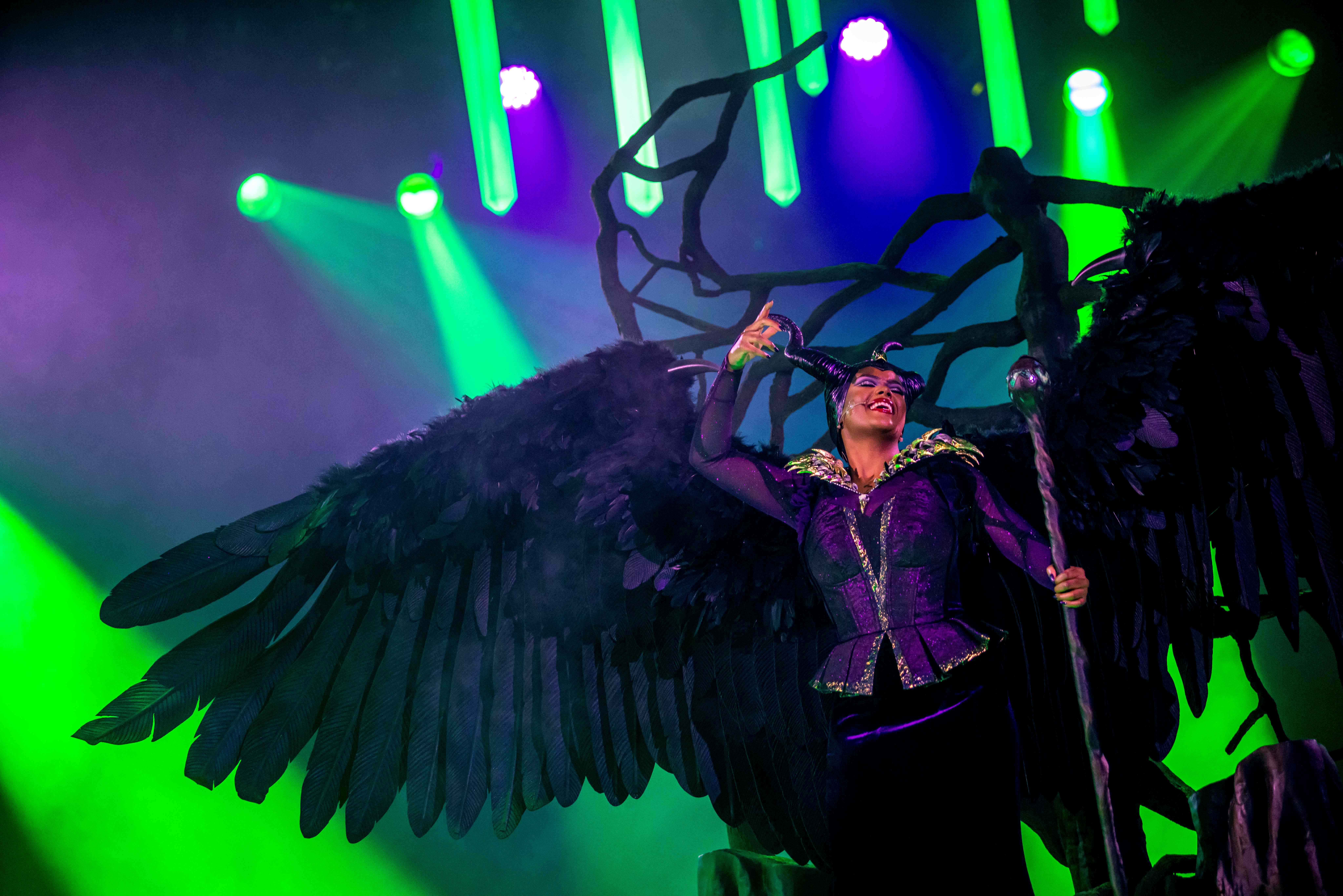 A performer in a dark, winged costume evokes Disney Villains on stage under dramatic green and purple lights.