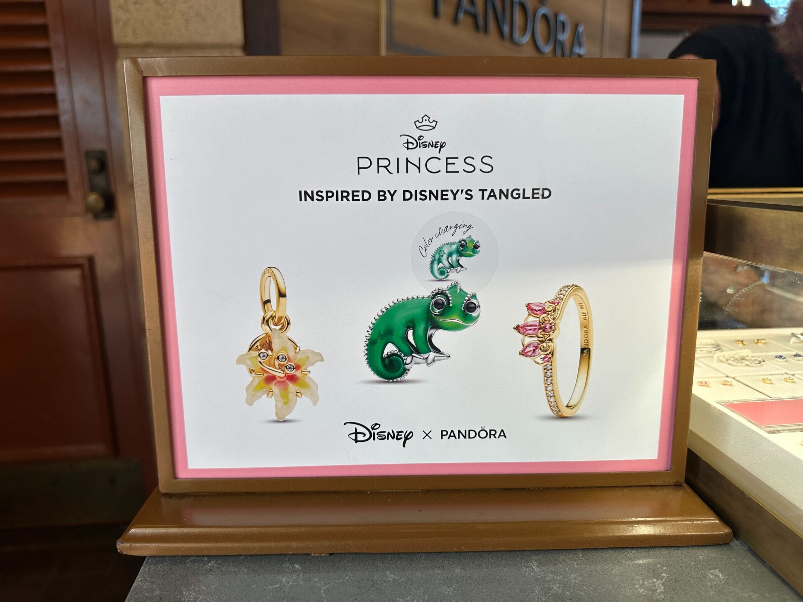 Display sign showing the items from the Pandora Tangled collection: a yellow sundrop flower charm, a green Pascal chameleon charm, and a gold tiara-shaped ring with pink stones.