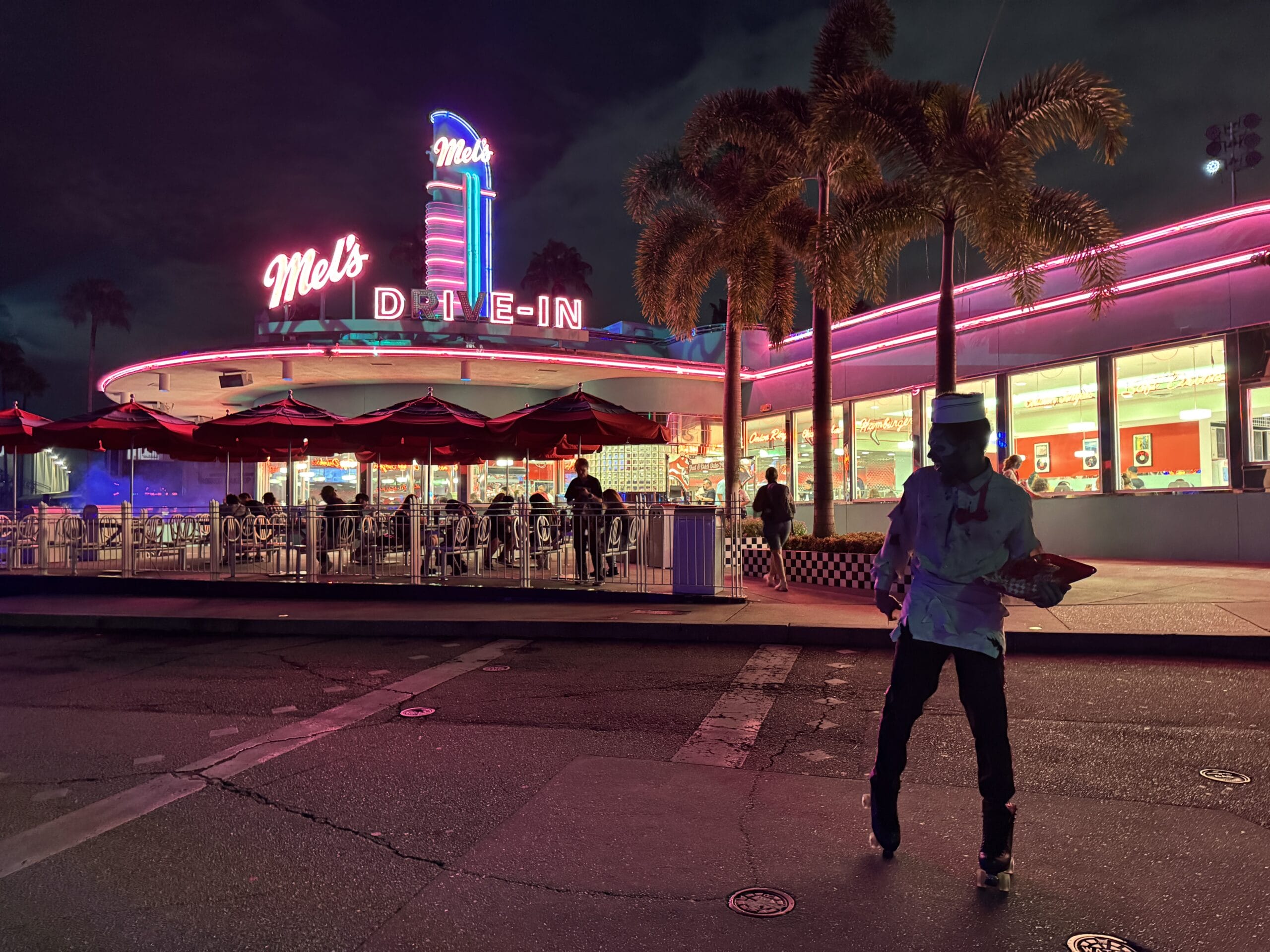 A neon-lit retro diner on Halloween Horror Night, with a server on roller skates and palm trees in the background.