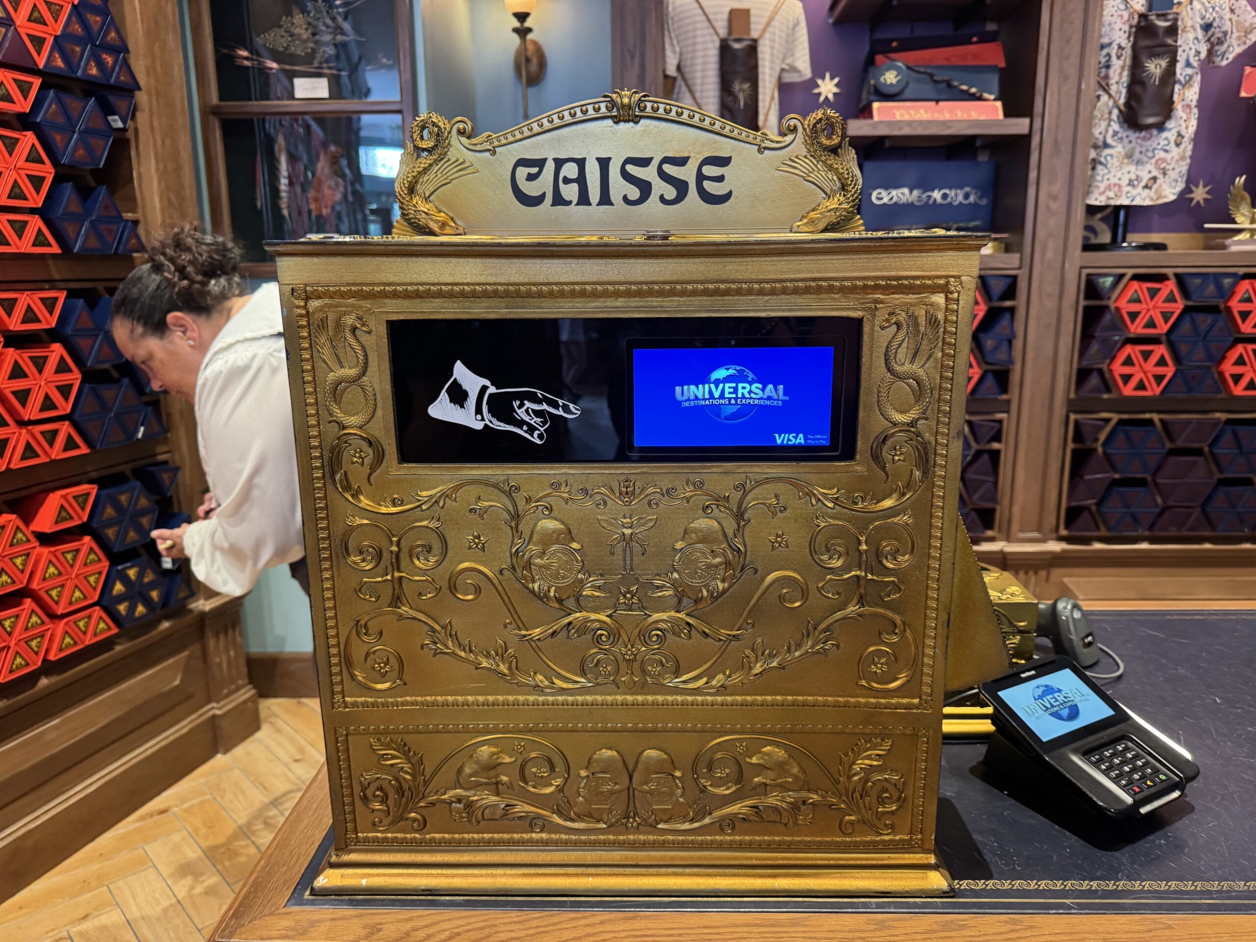 Ornate gold cash register labeled "Caisse," screen shows Universal, Visa, and Cosme Acajor Baguettes Magique logos.