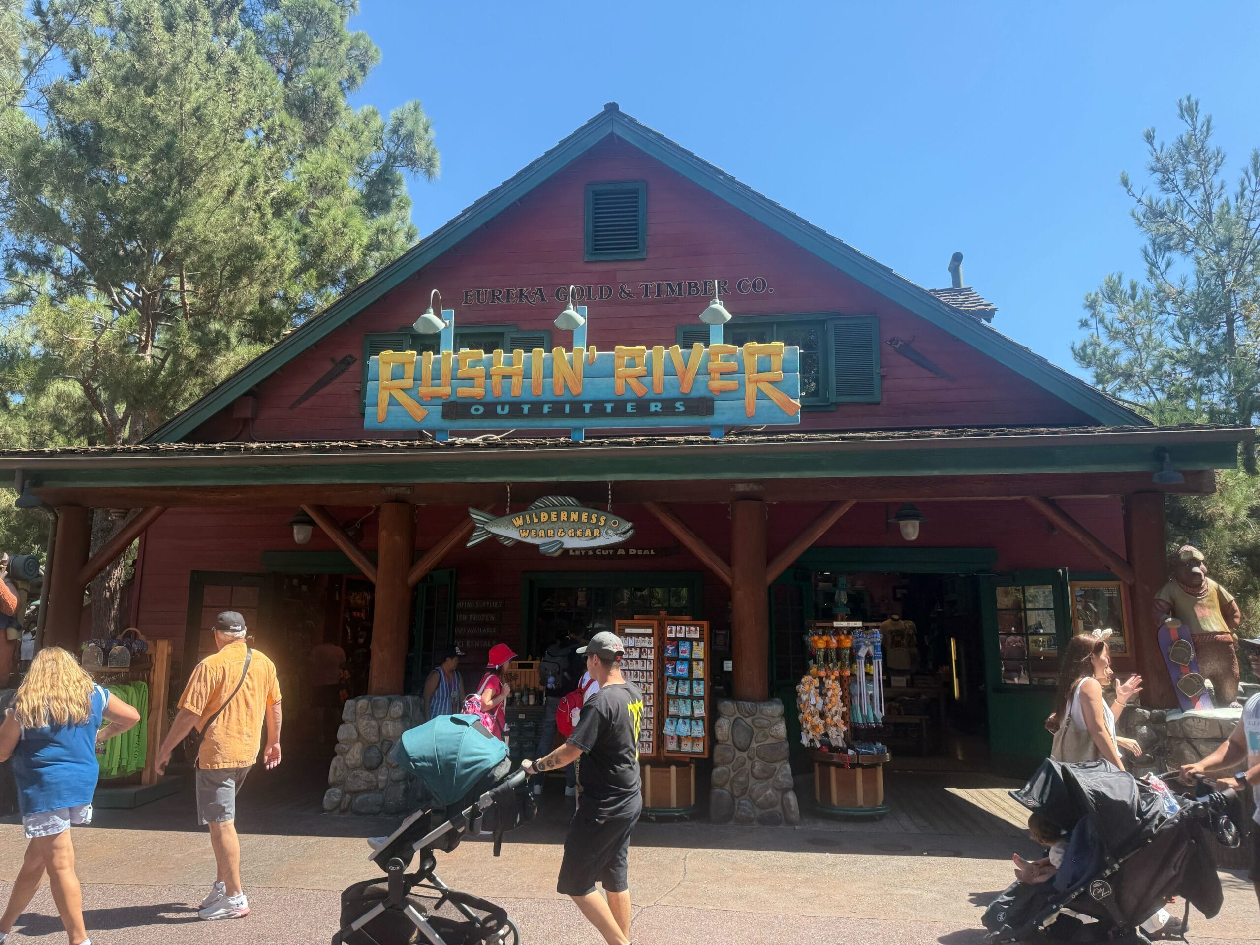 People walk past Rushin’ River Outfitters, a rustic, lodge-style gift shop with a green roof and wooden sign, on a sunny day.