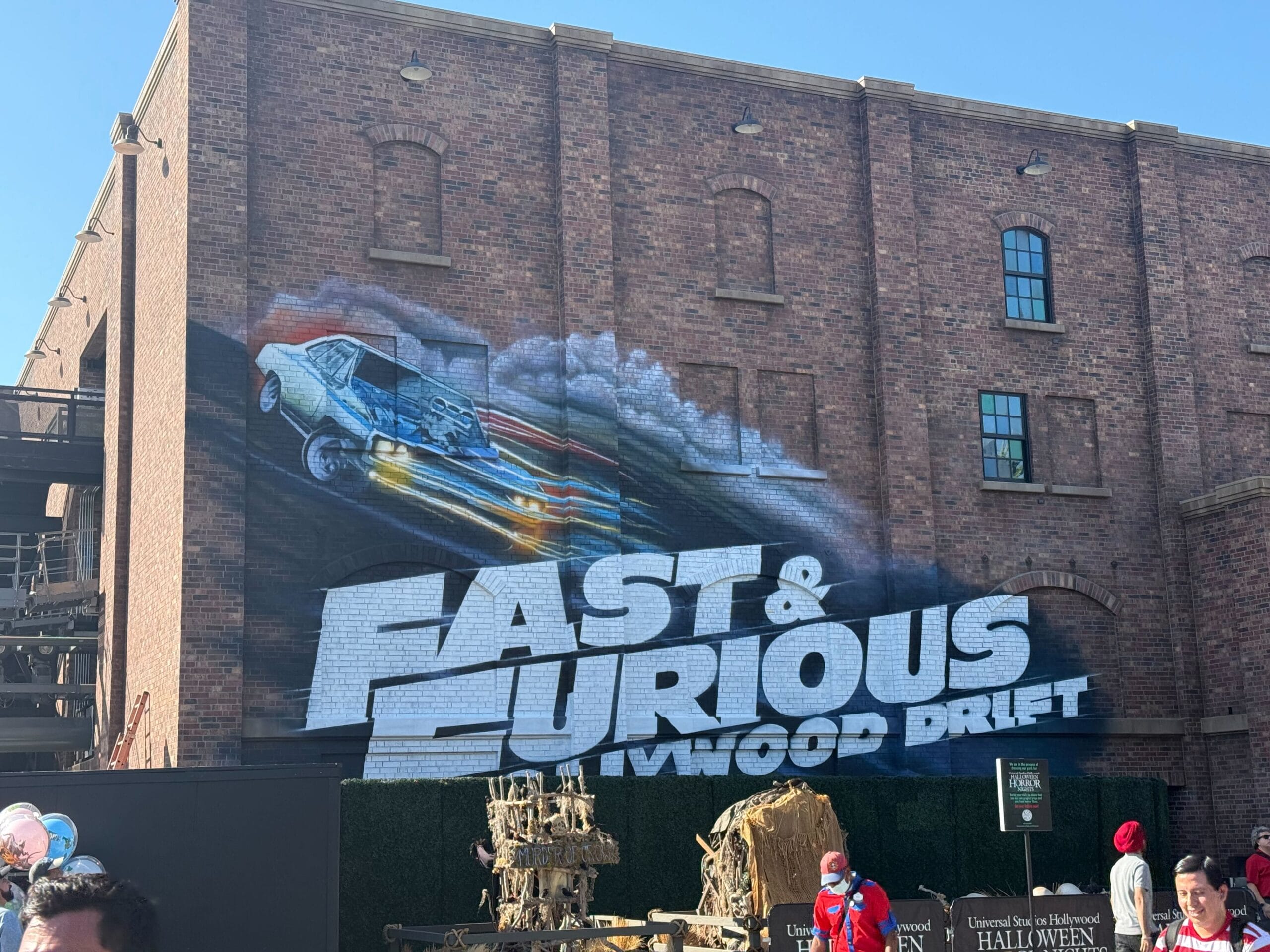A mural of a speeding car and "Fast & Furious: Hollywood Drift" on a brick building, with people walking by below.