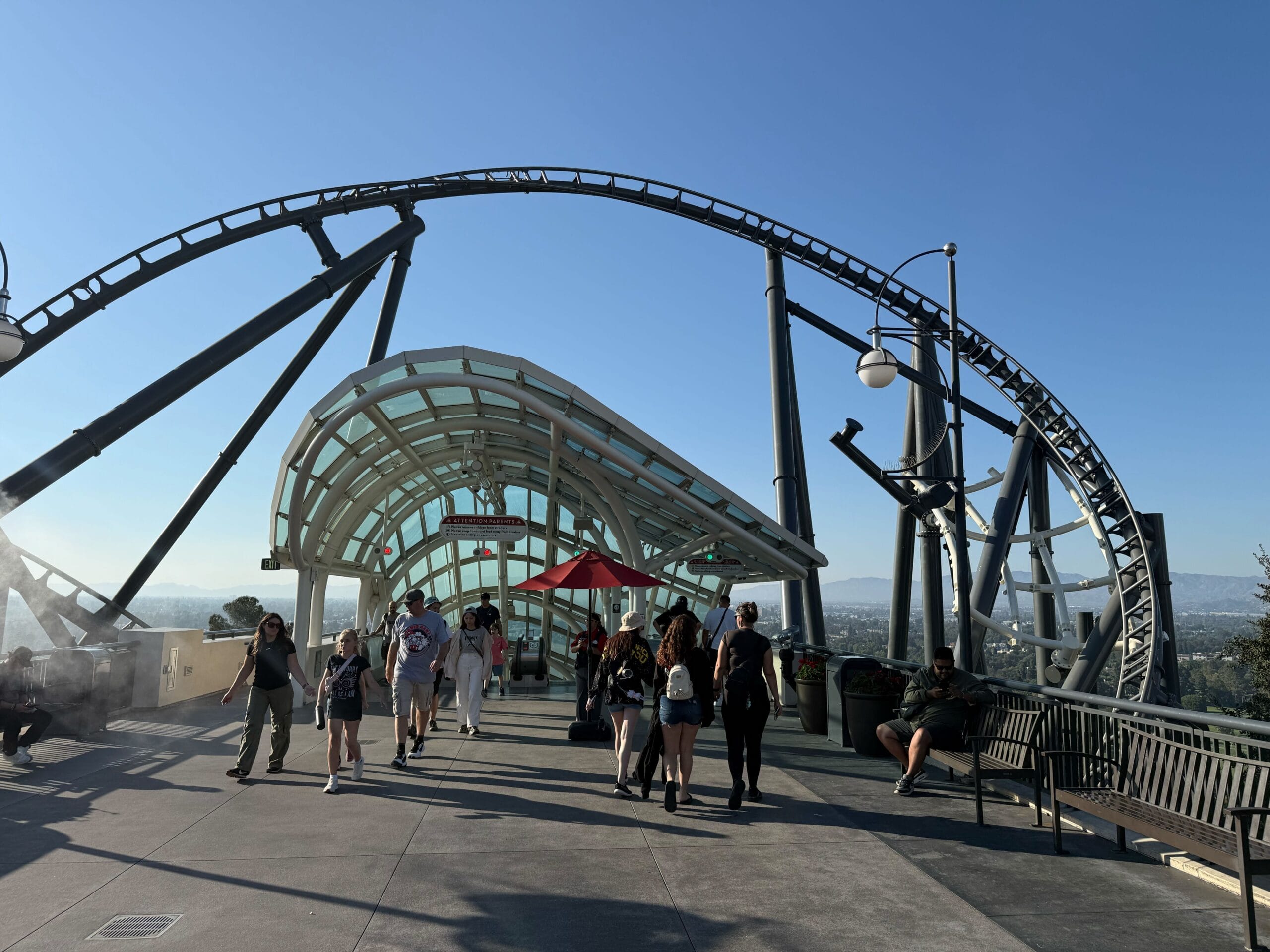 People walk under the Fast & Furious: Hollywood Drift roller coaster track near the glass escalator canopy on the Upper Lot at Universal Studios Hollywood.