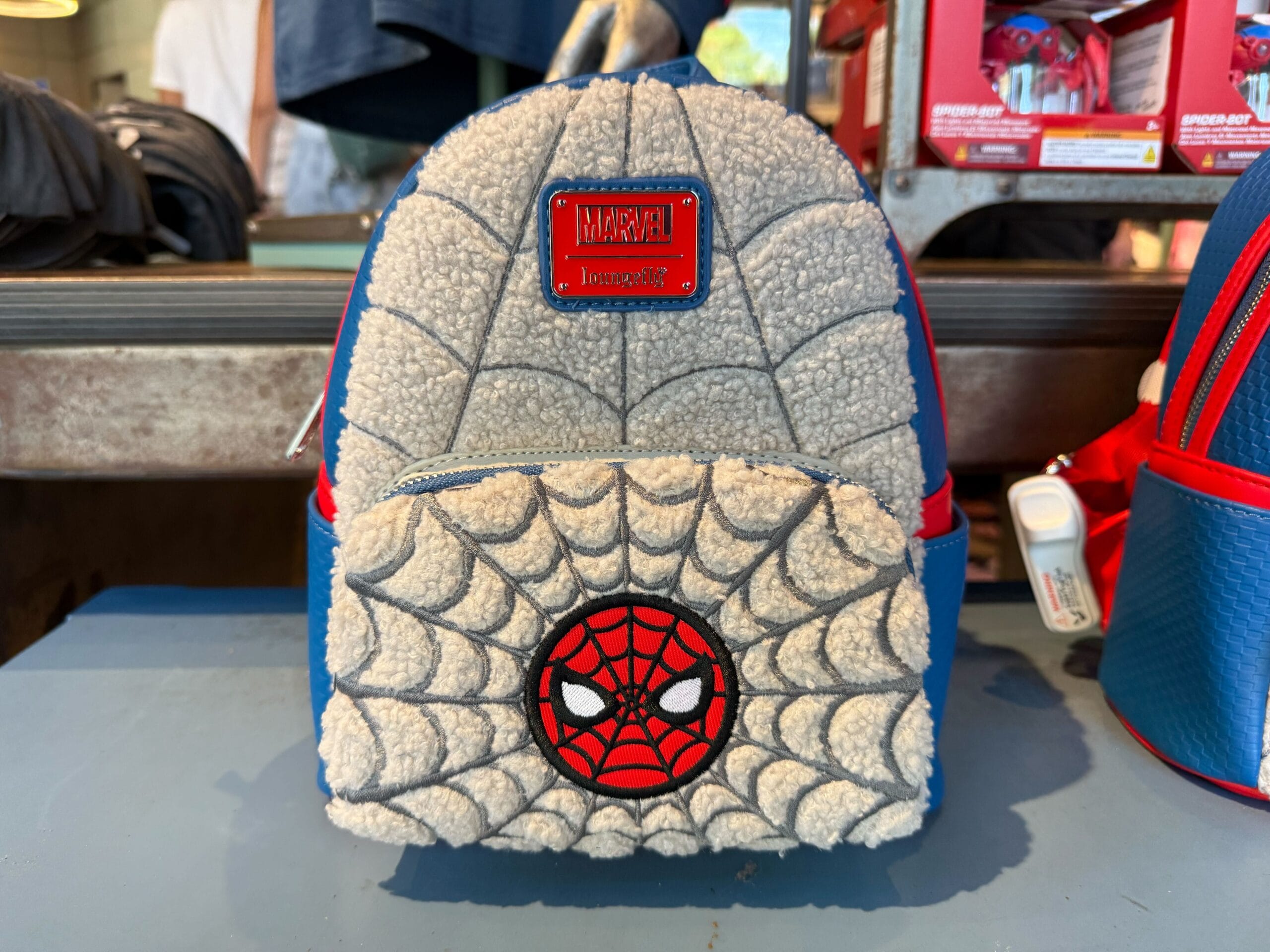 A fuzzy gray backpack with a web design and a red Spider-Man face on the front pocket, displayed on a store shelf.