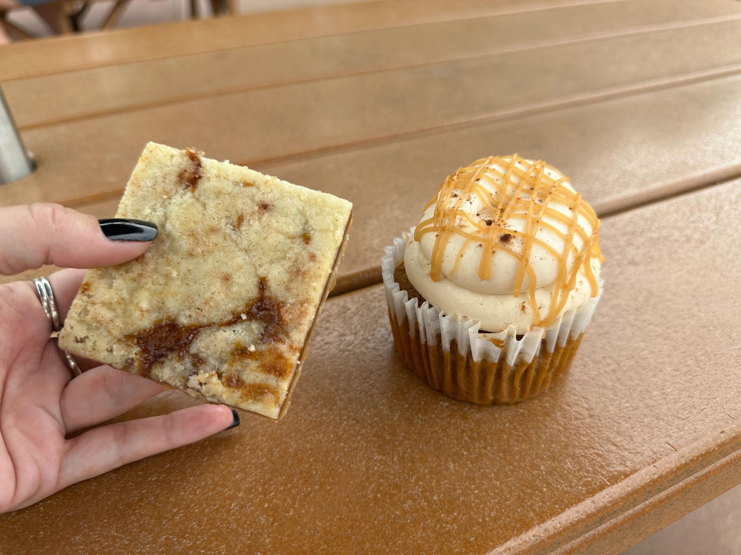 A hand holds a cinnamon streusel square beside a Werther's Original caramel-drizzled cupcake on a brown table.