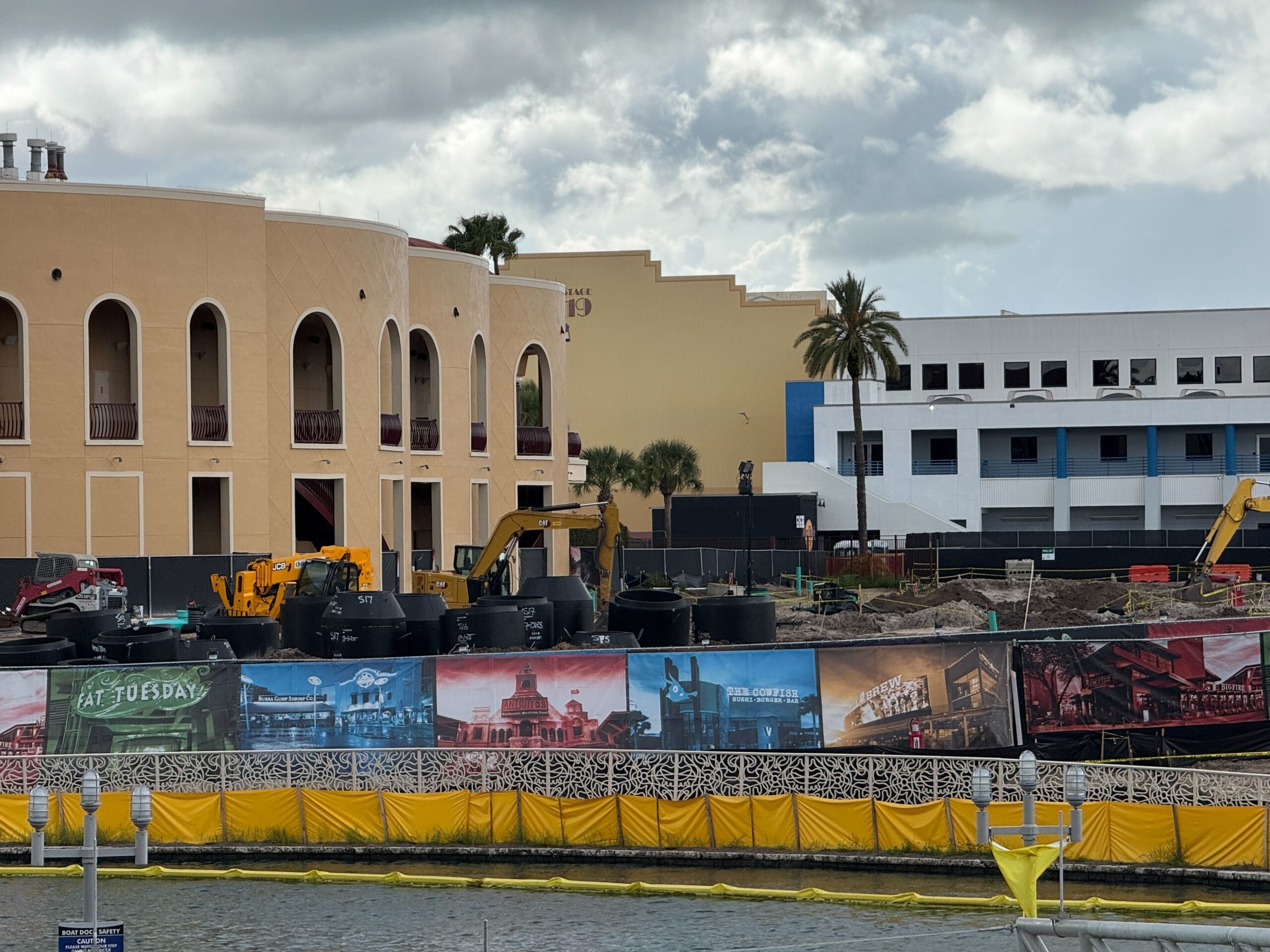 Construction site with excavators in front of a tan building, a palm tree, and colorful mural fence by water.