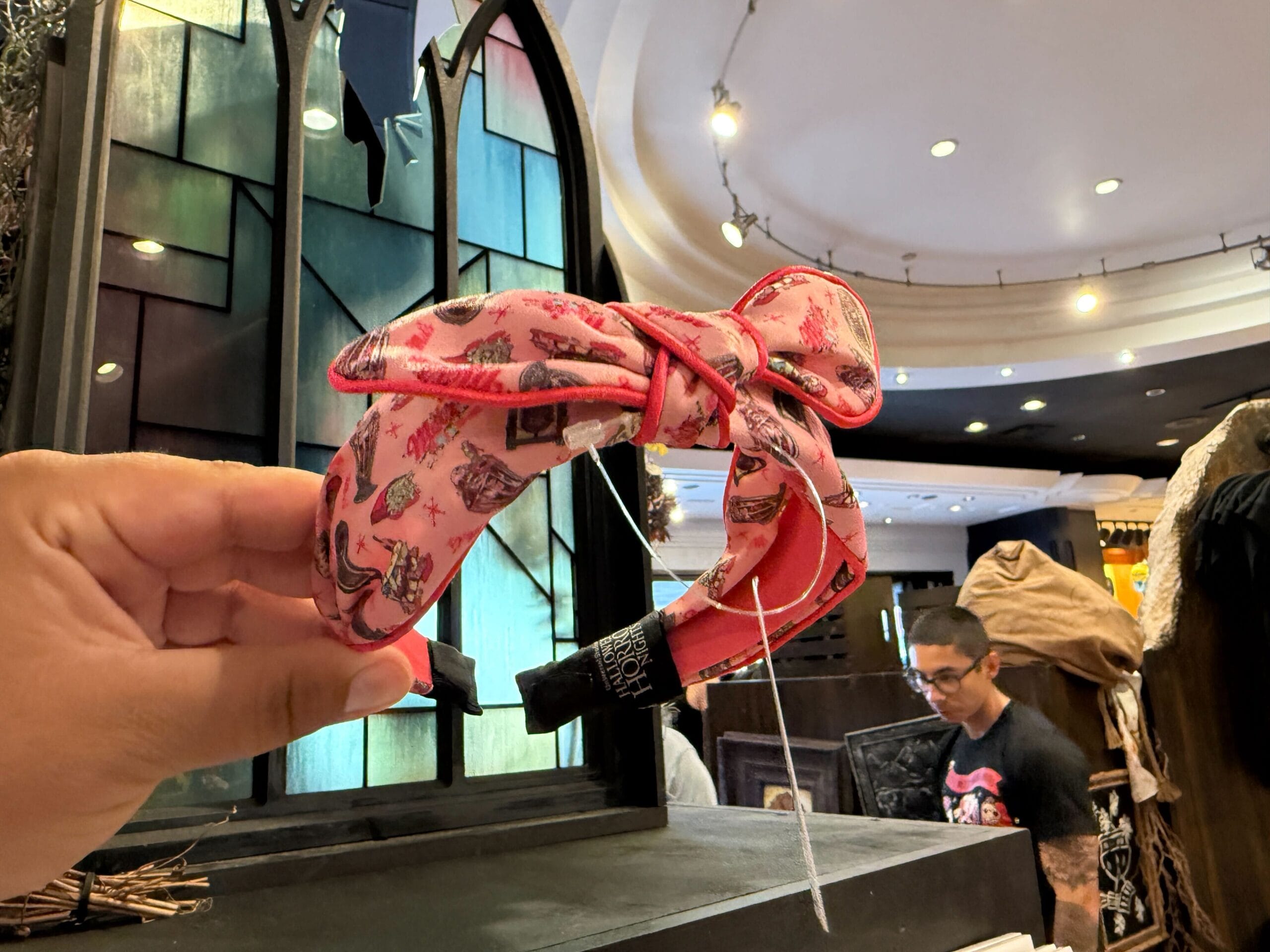 A hand holds a pink headband with a bow and Disney designs in a store with stained glass and people in the background.