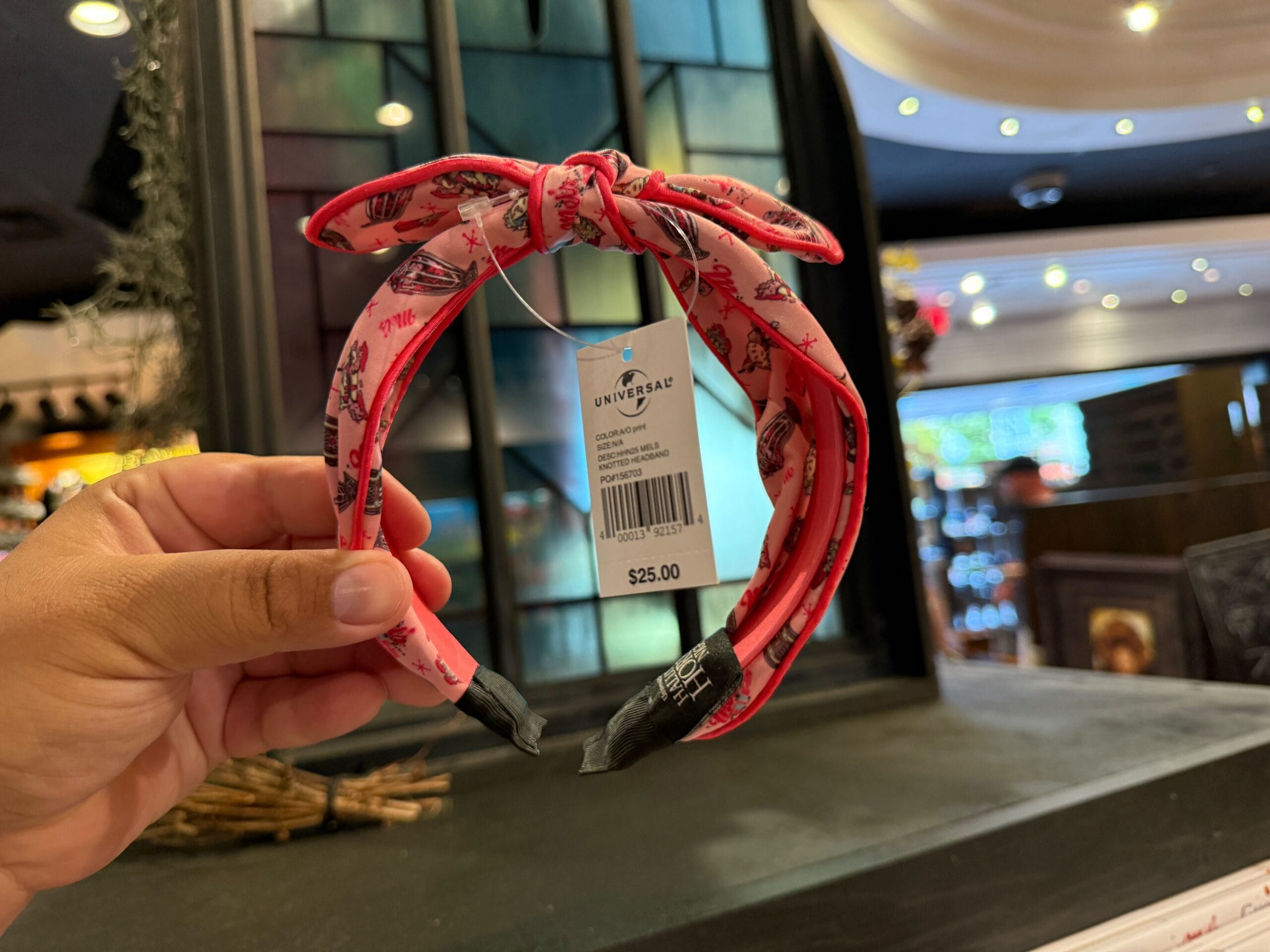 A hand holds a pink headband with a bow and cartoon print, showing a $25 price tag in a store.