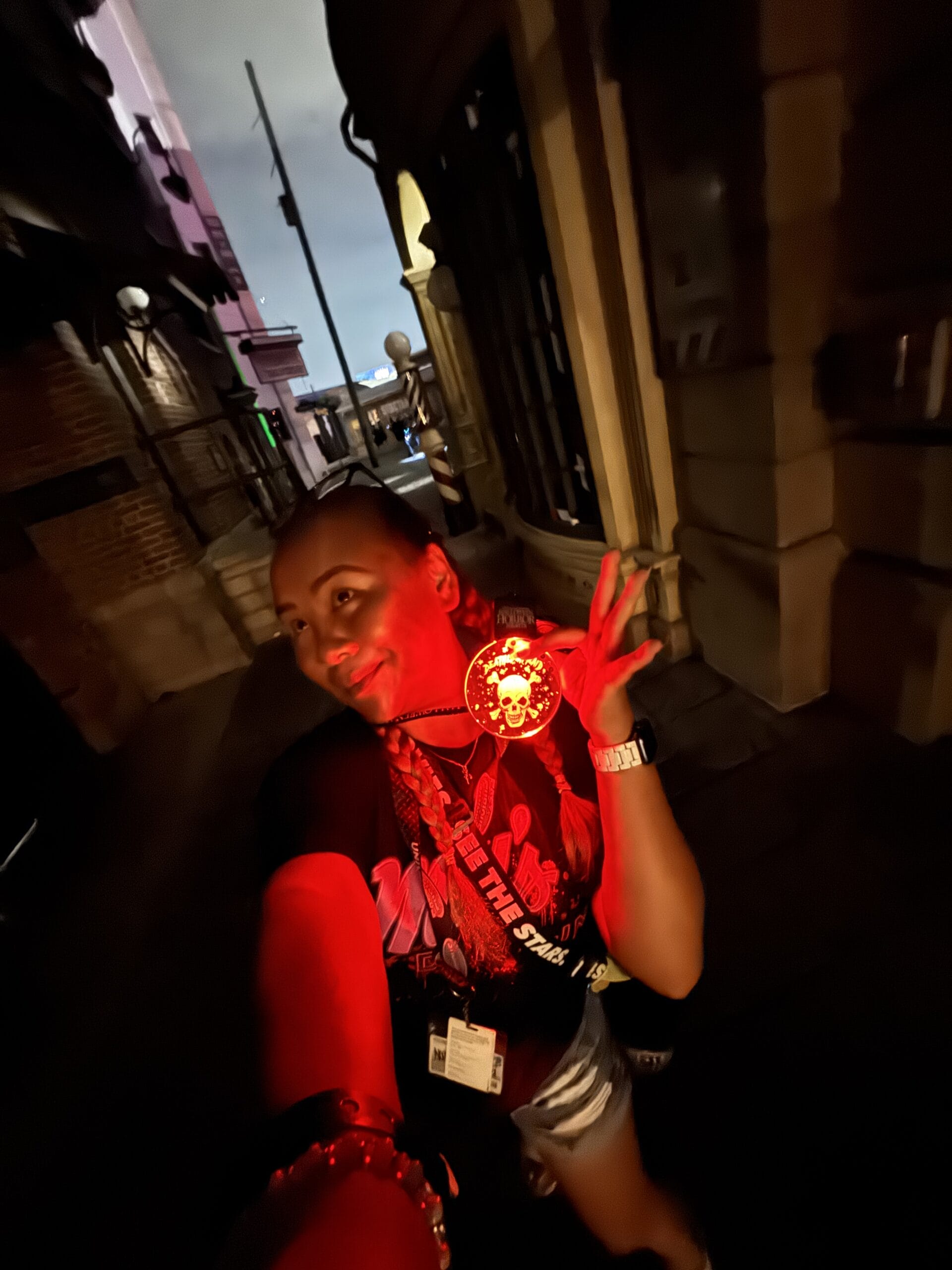 Person smiling at night, holding a red glowing medallion during Halloween Horror Night, on a dimly lit street with buildings.