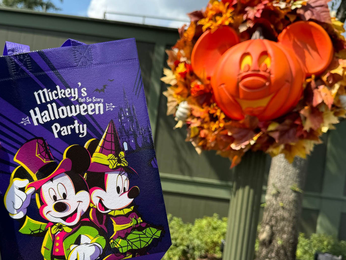 A purple Mickey’s Halloween Party bag is held near a Mickey-shaped pumpkin wreath decorated with autumn leaves.