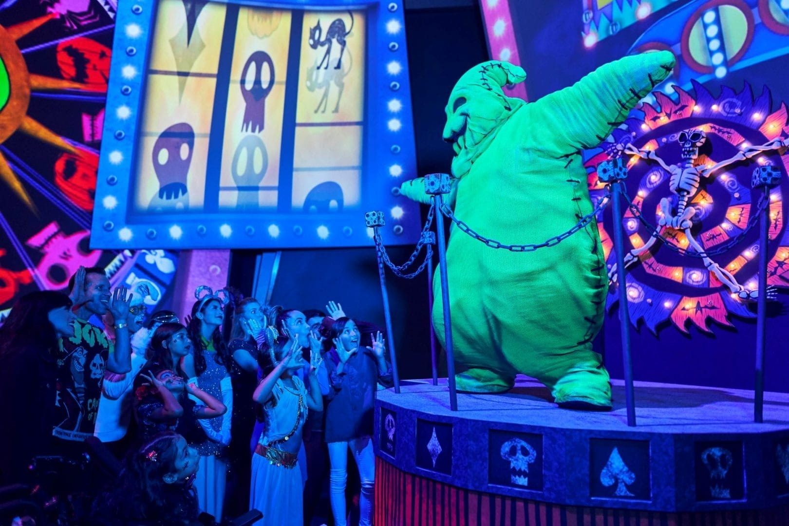 A crowd watches a large green character perform on a spooky, neon-lit stage with Halloween-themed decorations.
