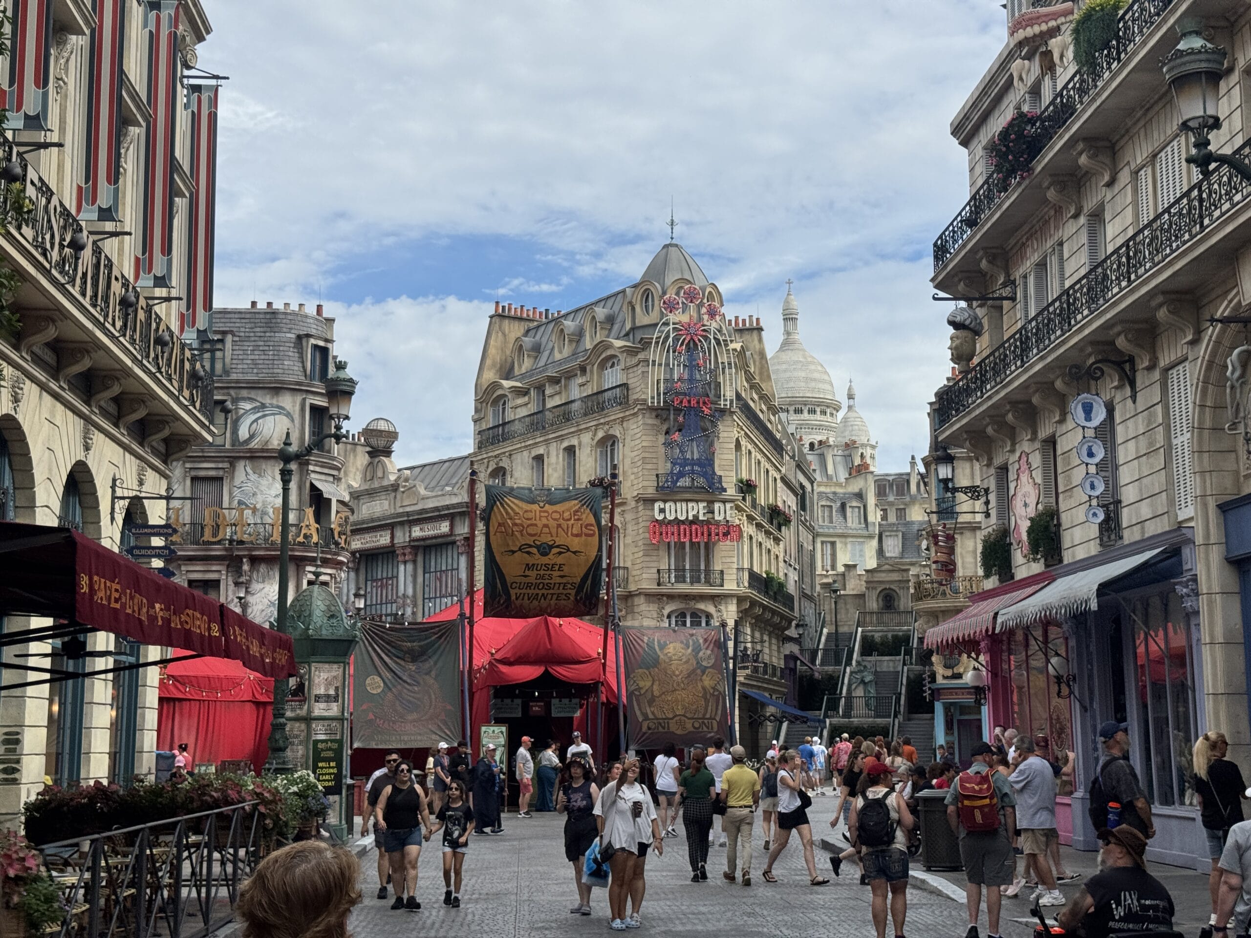 Crowds stroll an Epic Universe street with ornate buildings and a domed church under a clear blue sky.