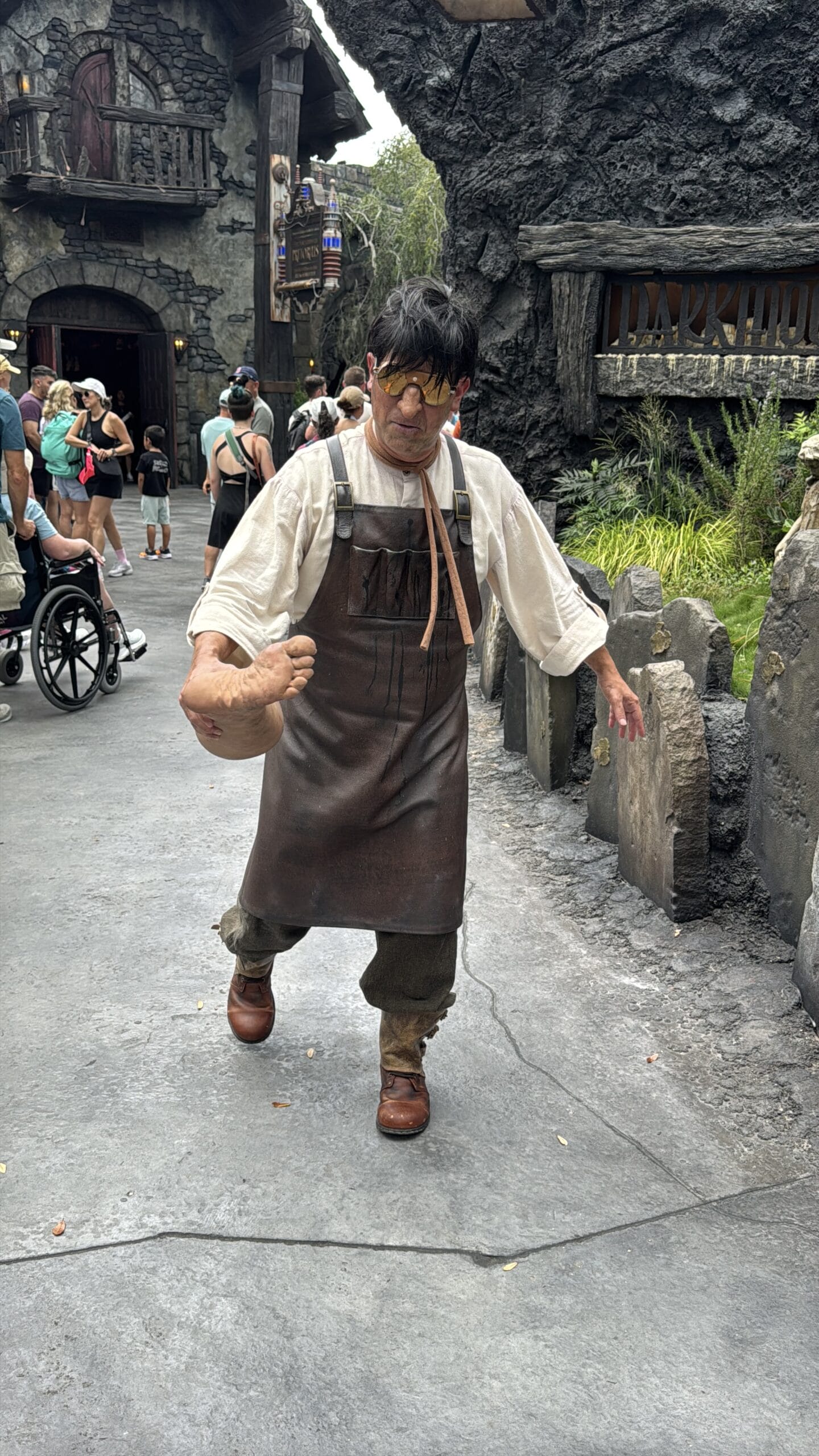 A person in a brown apron and gloves acts as a character with a fake arm at Epic Universe, entertaining park guests nearby.